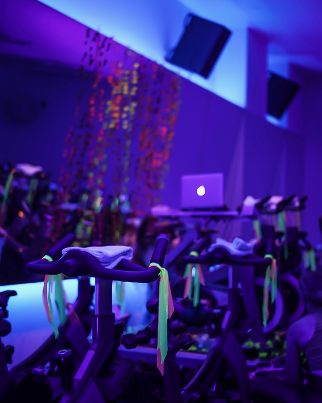 Indoor event with exercise bikes, a laptop on a table, and colorful lighting, possibly a fitness or dance class.