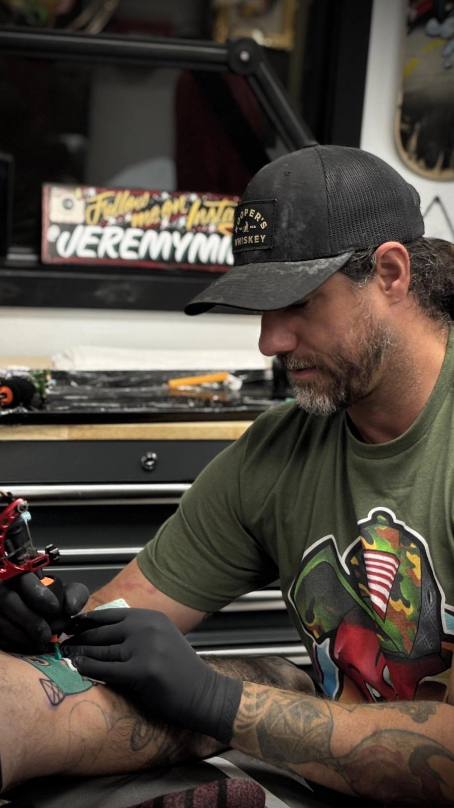 Man with a beard wearing a black cap and green T-shirt tattooing a person's arm in a tattoo studio.