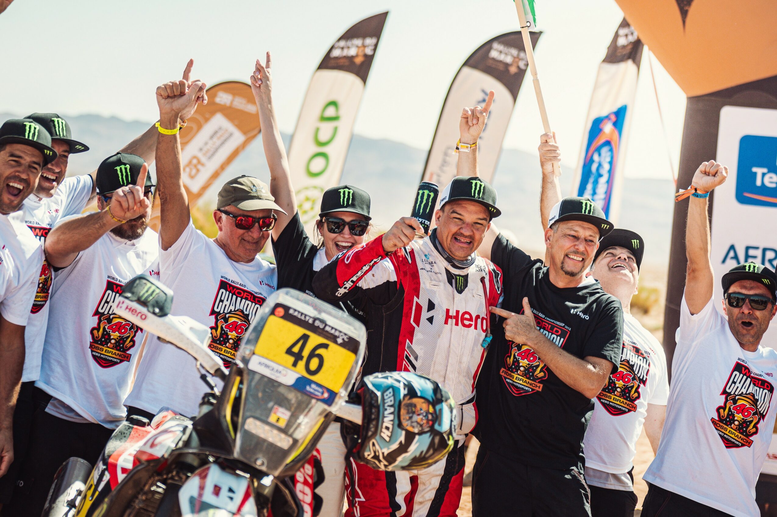 Group of people celebrating at a racing event, some wearing Monster Energy hats and racing gear, holding flags and raising fists.