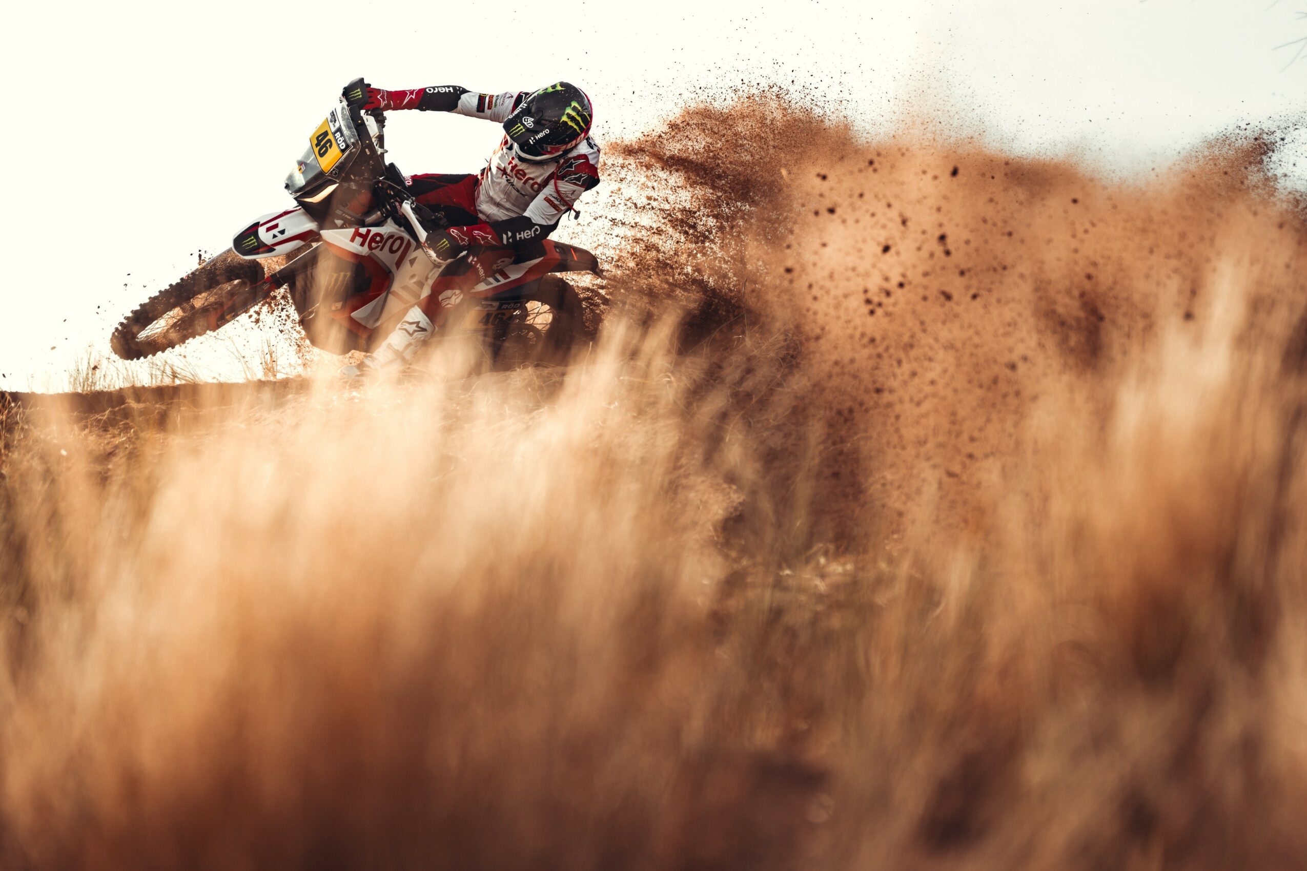 Motorcycle rider in mid-air, kicking up dirt on a dry, grassy terrain, with a white sky background.