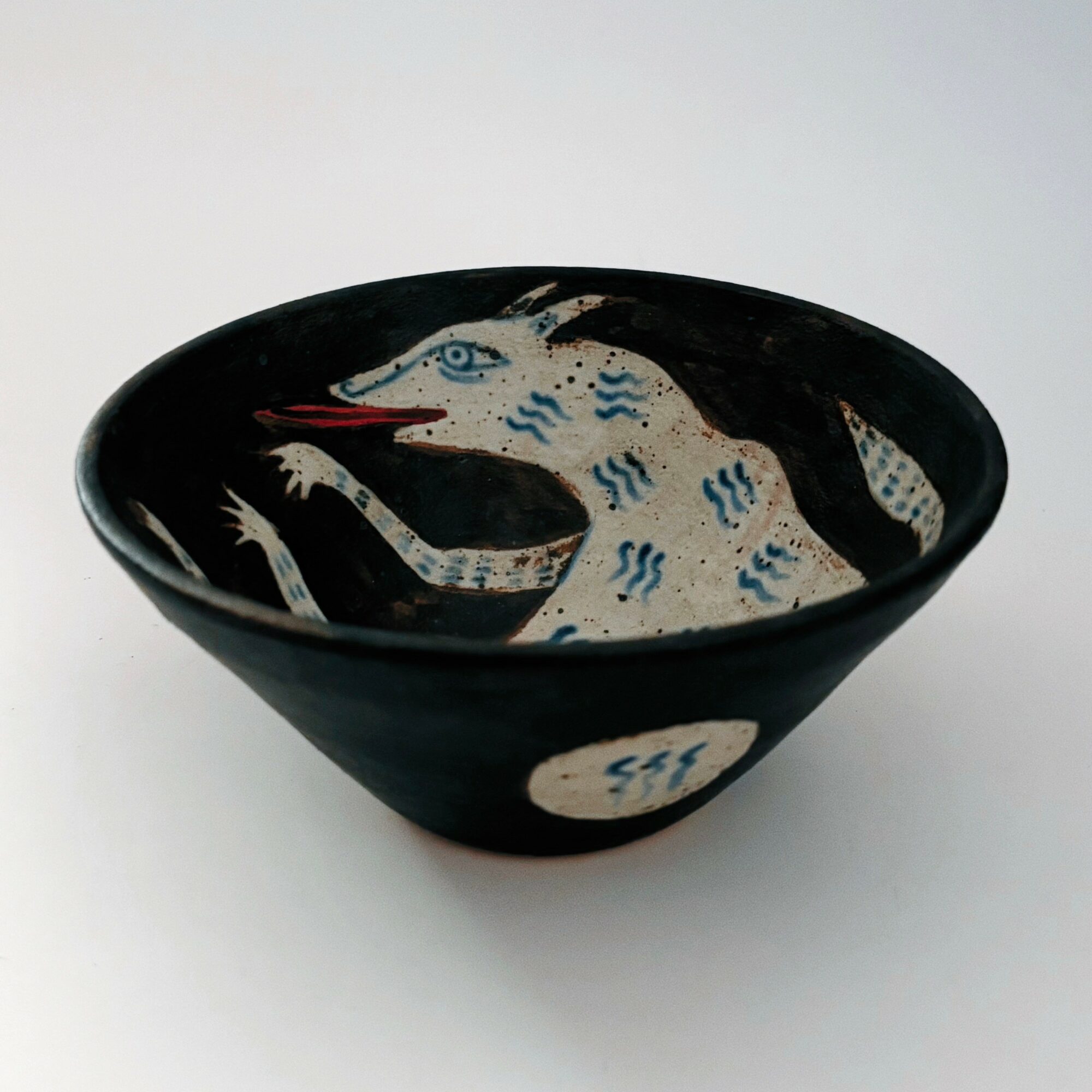 Black bowl with a painted white and blue stylized animal figure, possibly a dog or wolf, on a white background.