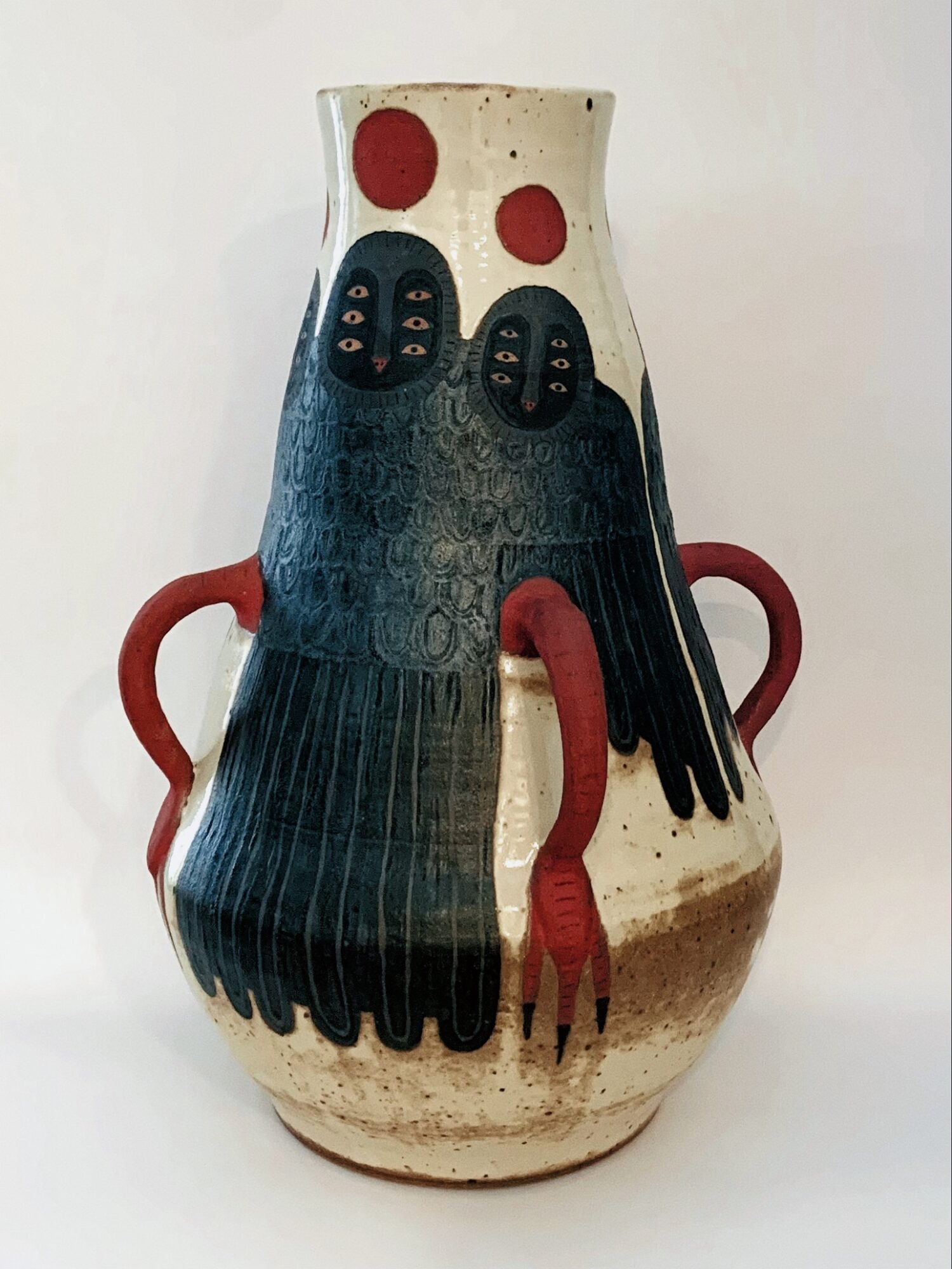 Decorative ceramic vase with abstract face design, red handles, and red circles on top, against a plain background.