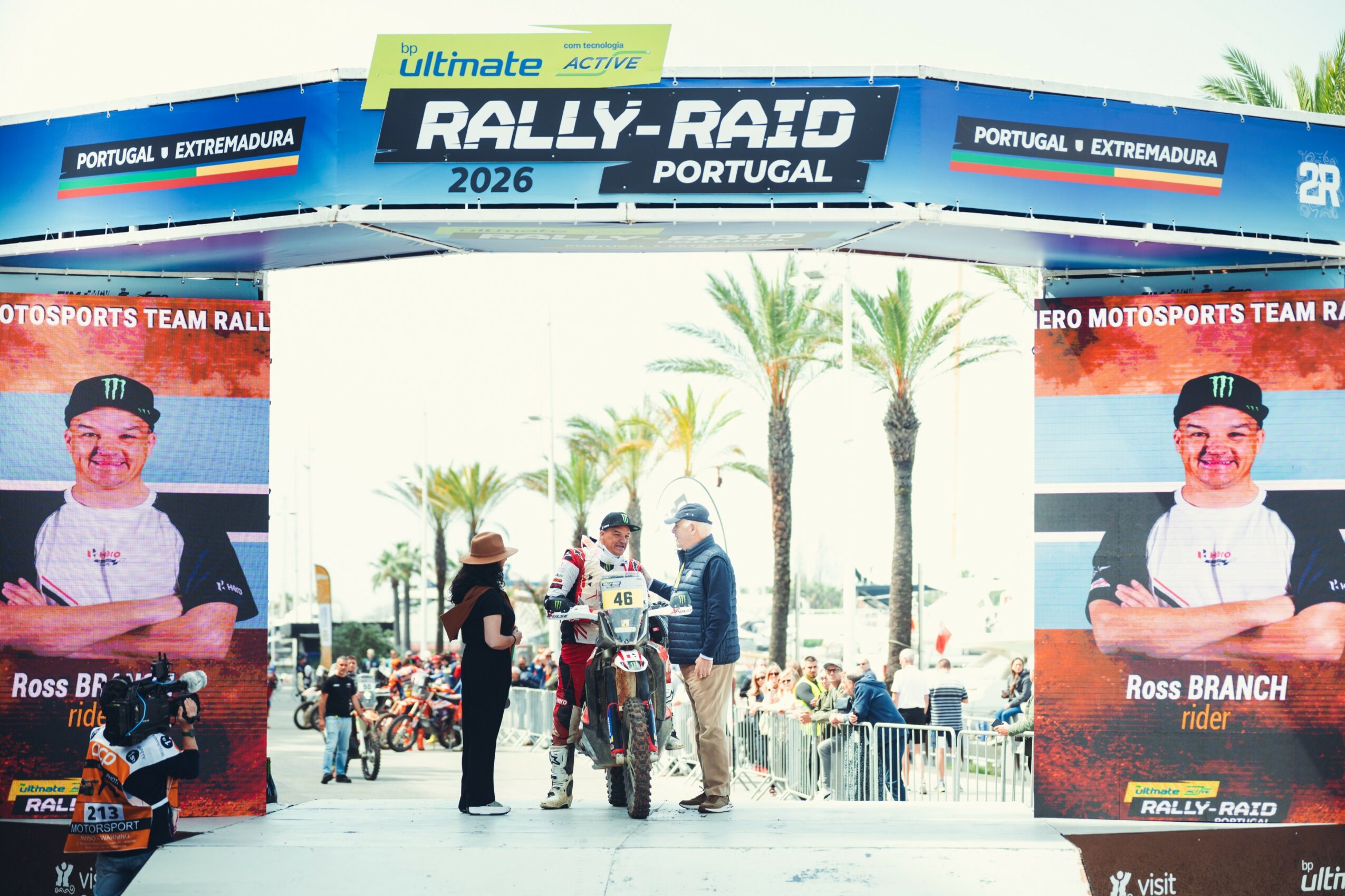 Rally event entrance with banners, palm trees, and people gathered, under a blue sky.