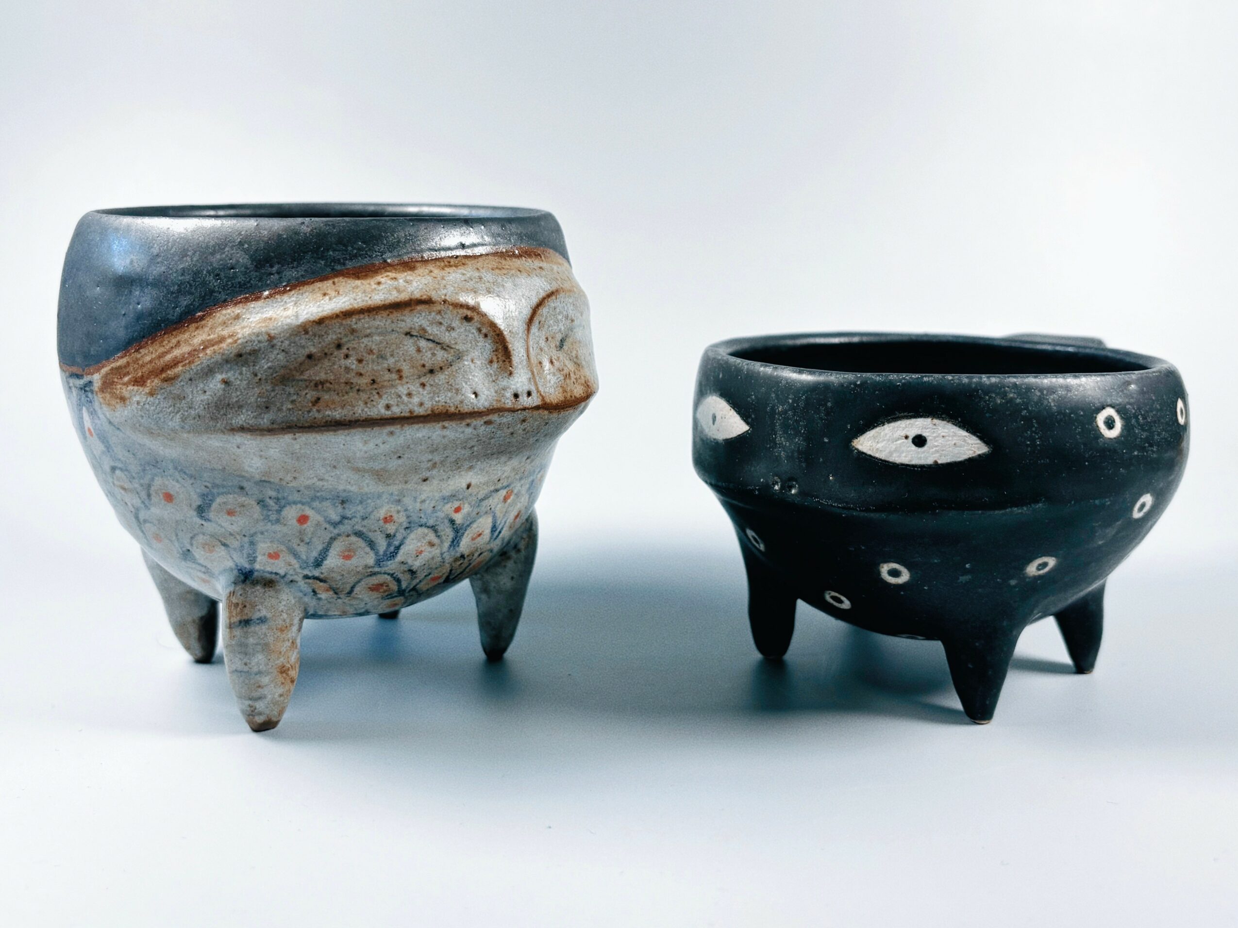 Two ceramic pots with three legs each, one with a blue and brown glaze, the other black with white eye-like patterns, on a white background.
