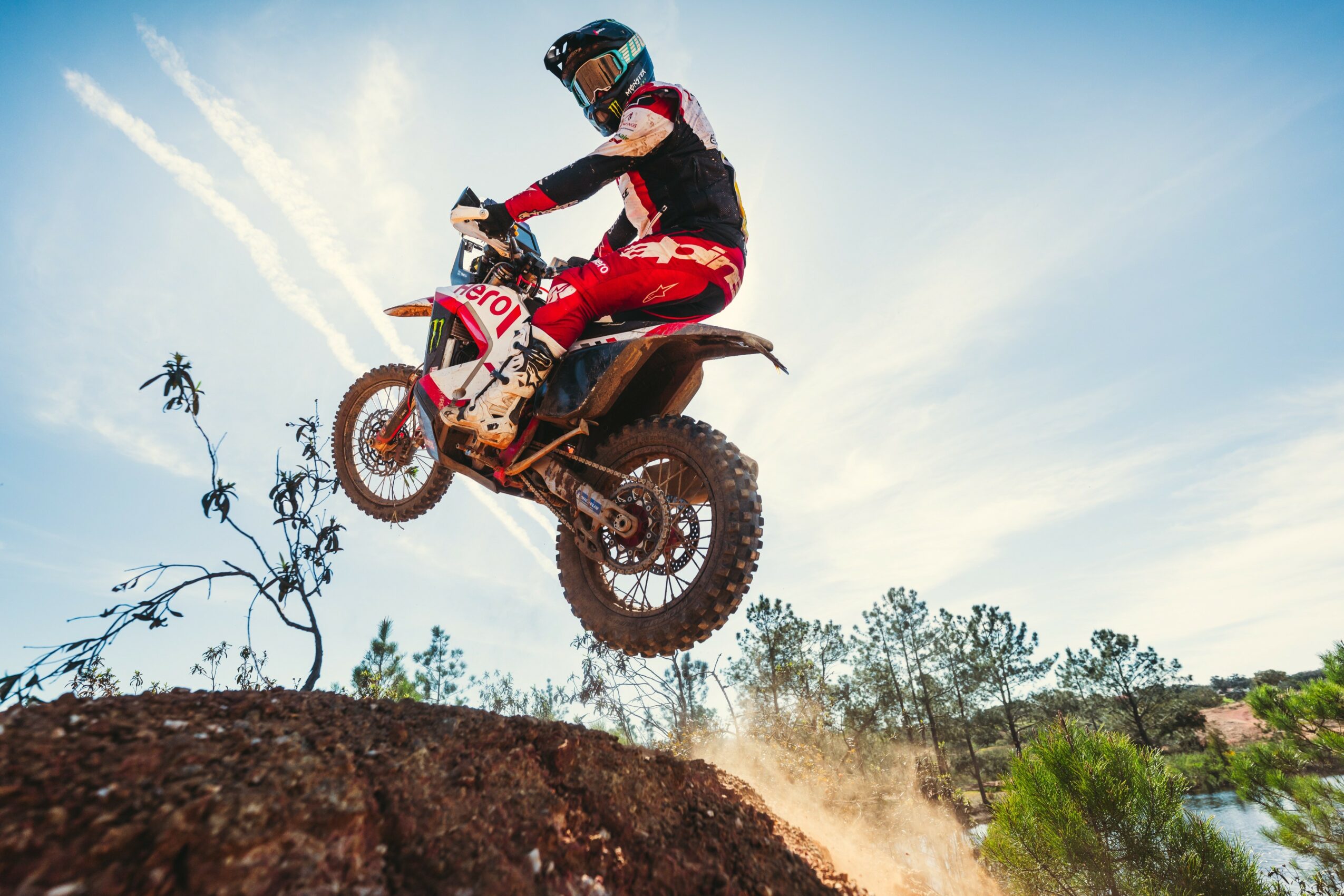 Motorcycle rider in mid-air jumping over dirt mound during daytime, with trees and sky in background.