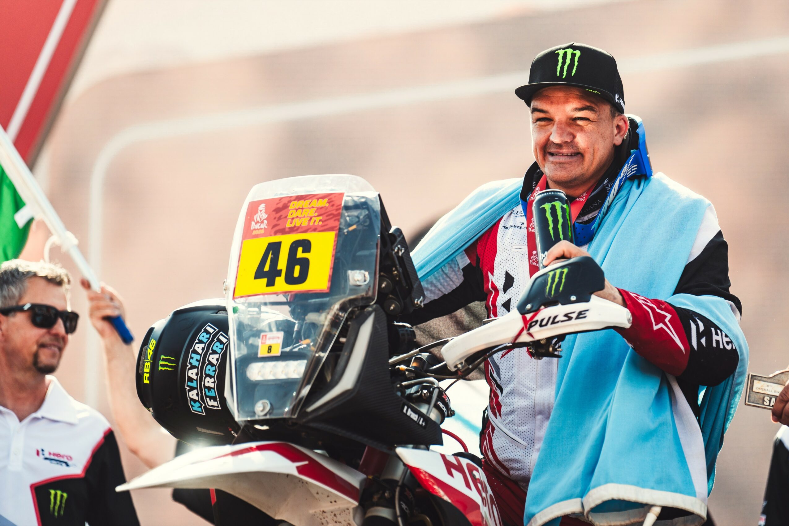 Man smiling at a motorcycle race, wearing a cap and racing gear, with a race number 46 on the bike, in a pit area.