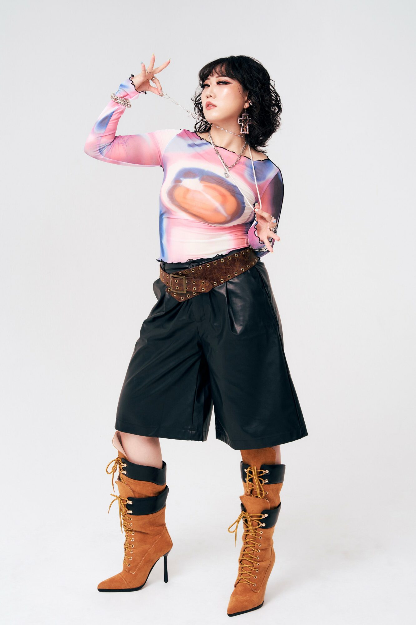 Woman with curly hair wearing a colorful top, black wide-leg shorts, and brown high-heeled boots, posing with her hand near her face.