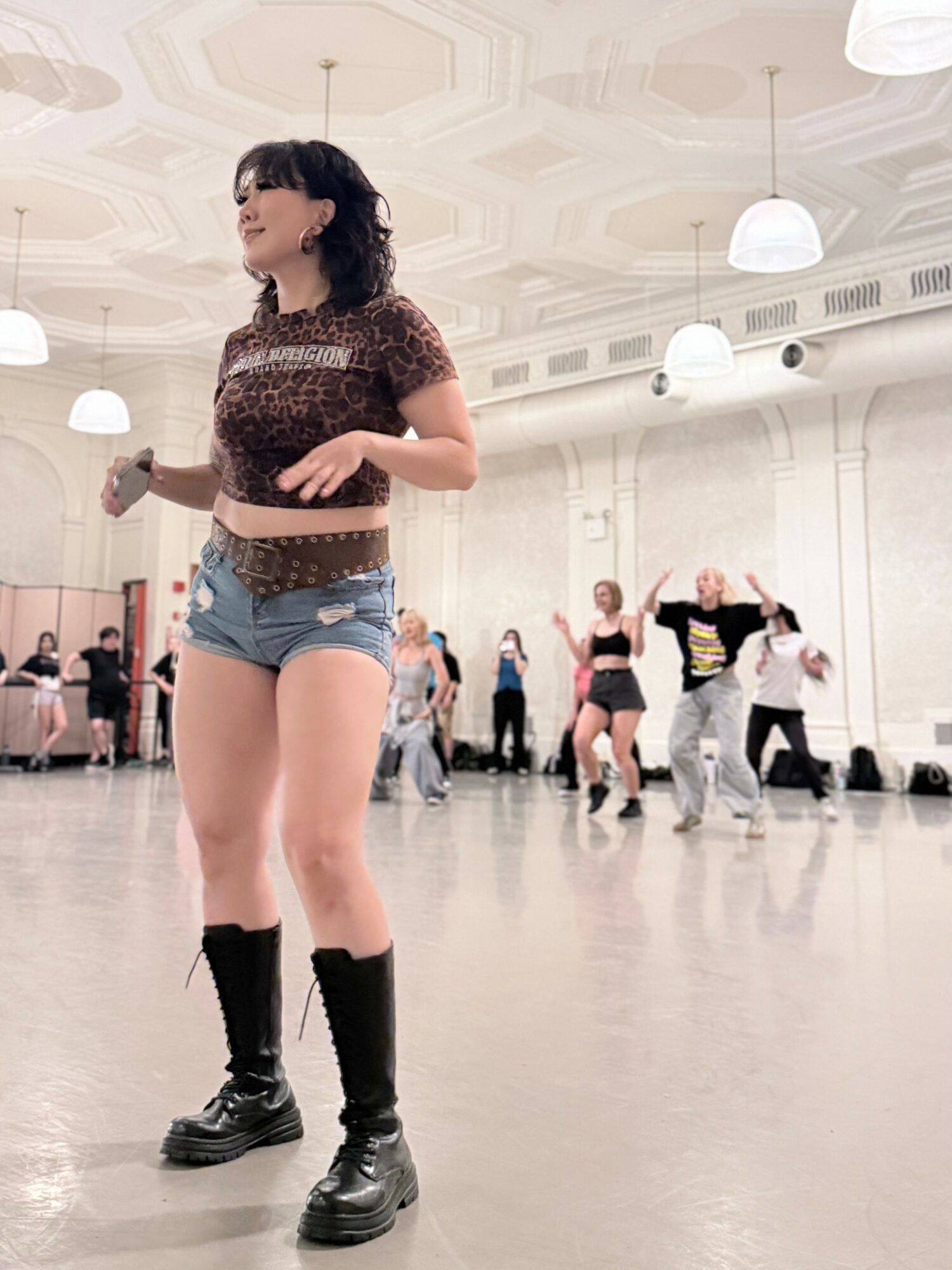 Young woman with curly hair in a dance studio, wearing a patterned crop top, denim shorts, and black boots, smiling.