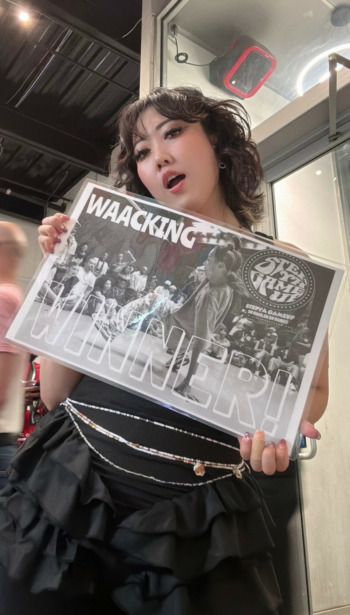Young woman with curly hair holding a black-and-white poster, standing indoors with a reflective surface behind her.
