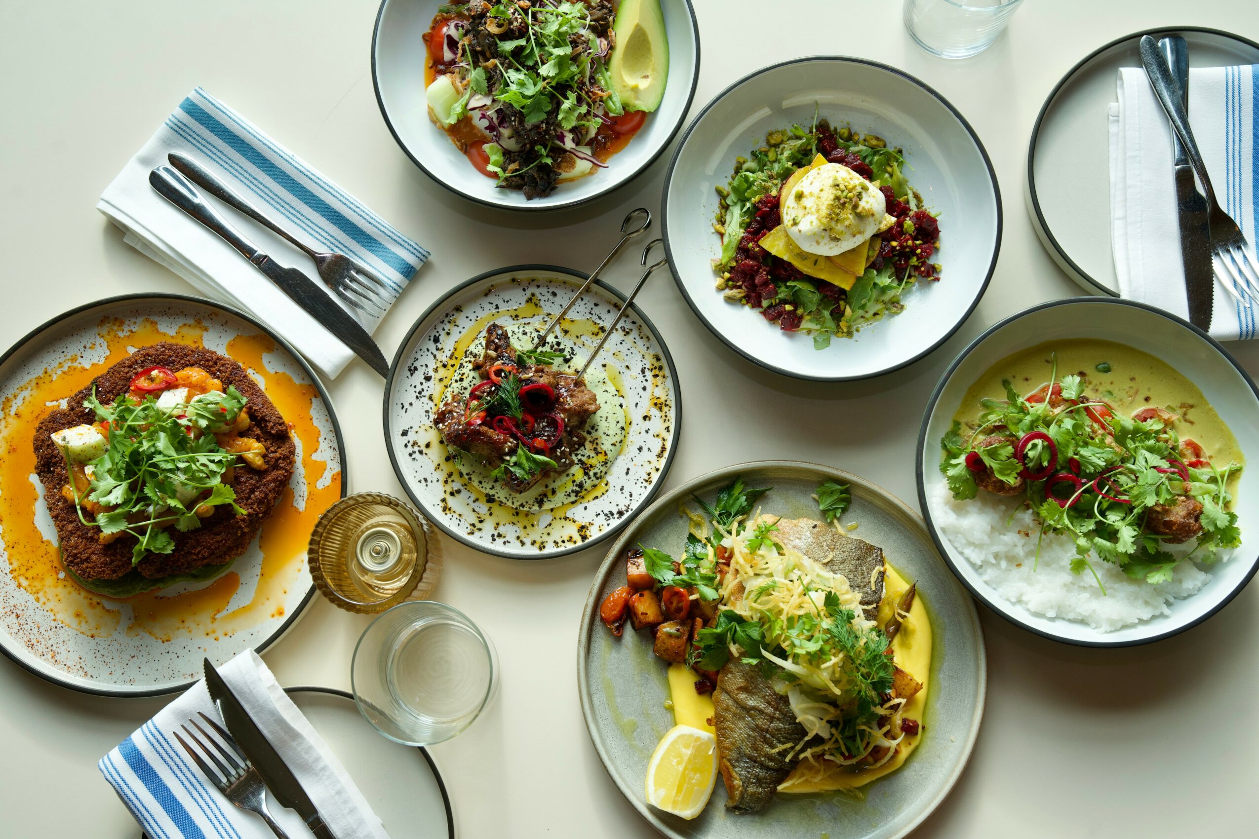 Table with various salads and dishes, cutlery, glasses, and plates arranged on a white surface.