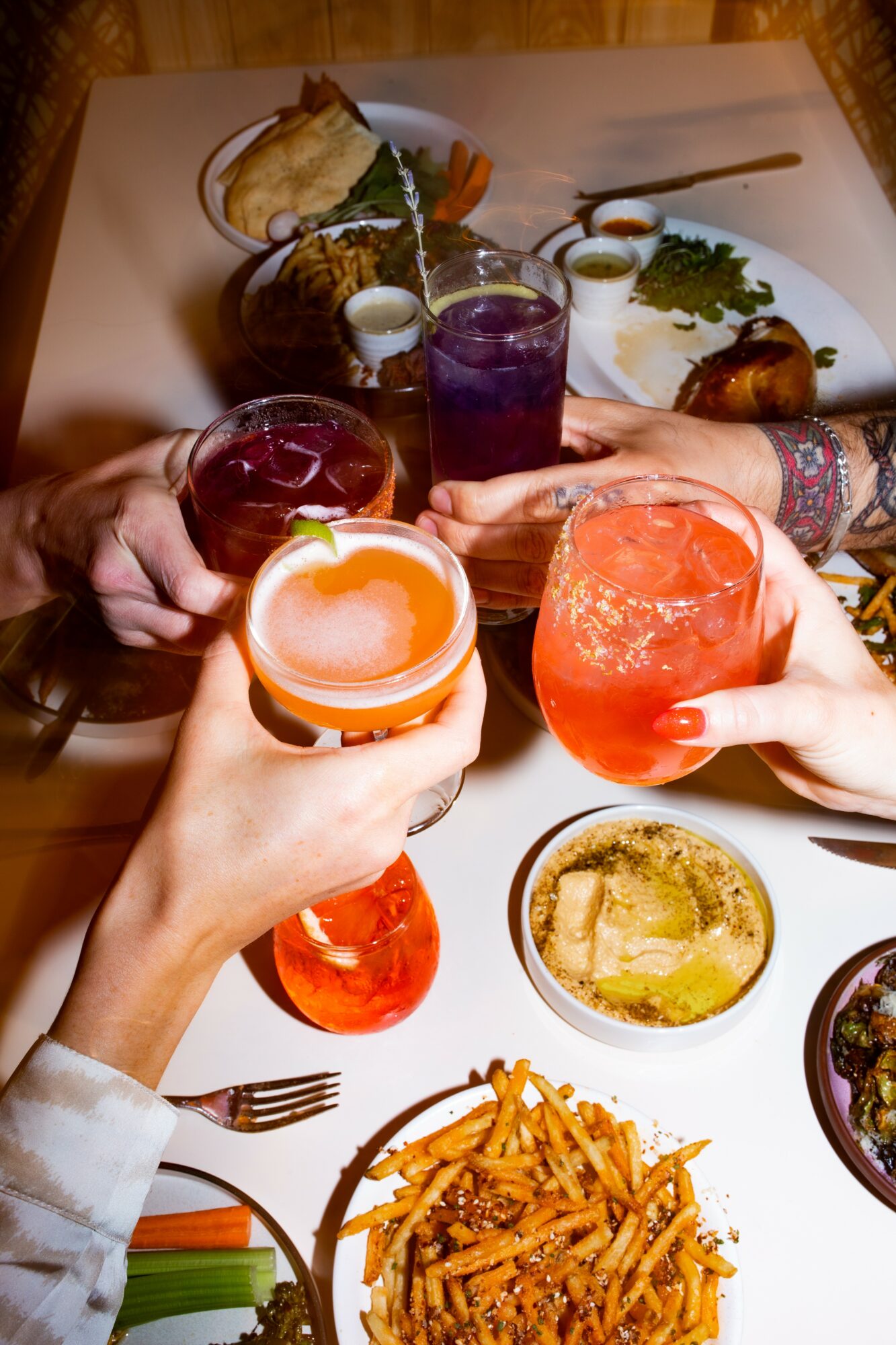 Four people hold glasses of colorful drinks over a table with food, including fries and dips, in a dining setting.