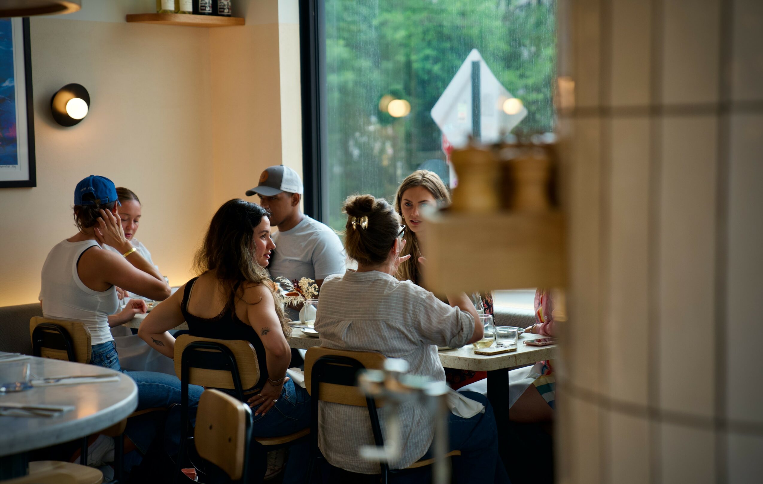 Group of people sitting at a table inside a cafe, with a window showing trees outside, seen from behind a wall.