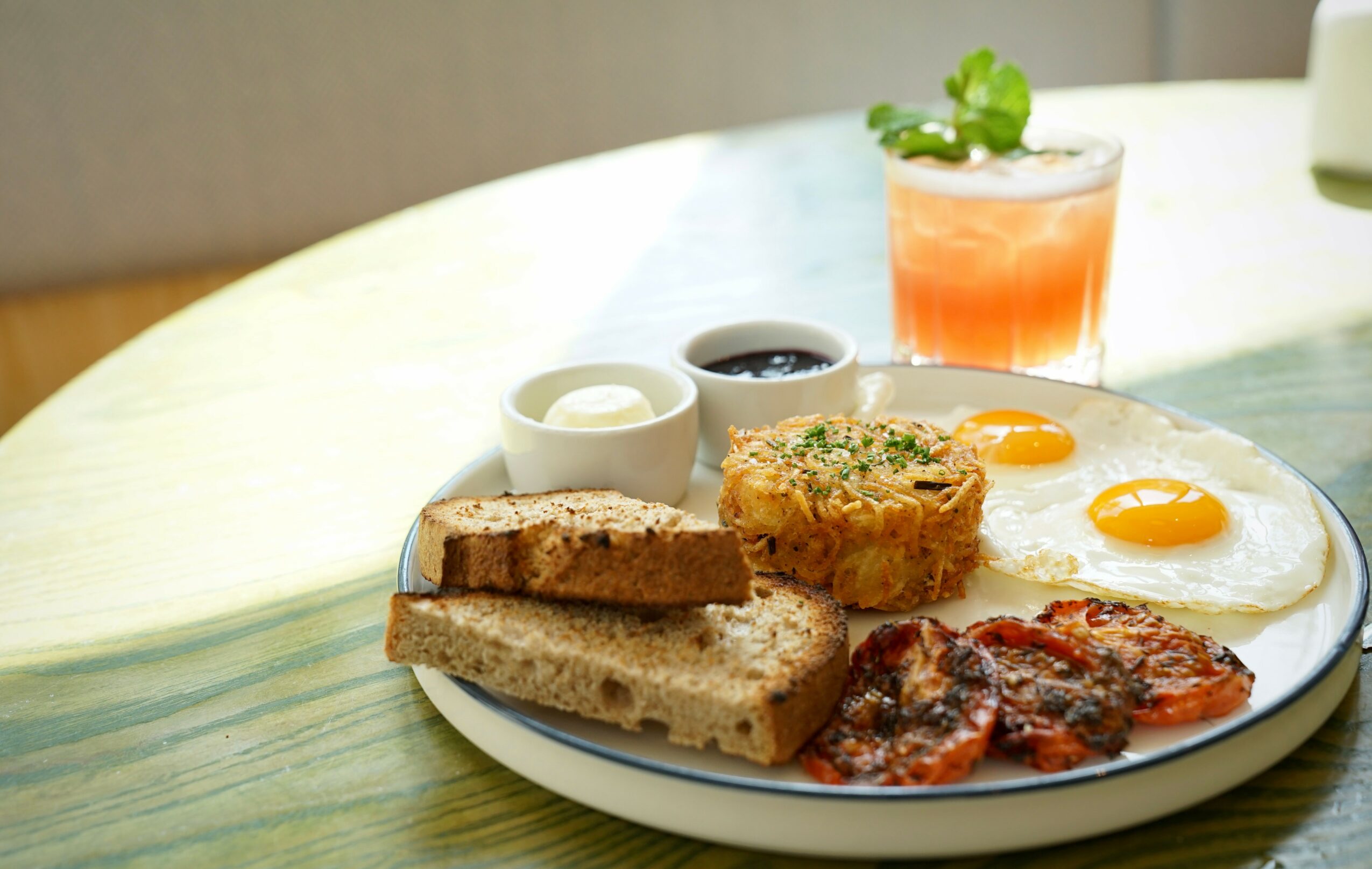 Breakfast plate with fried eggs, toast, bacon, and sides, on a table with a drink in the background.
