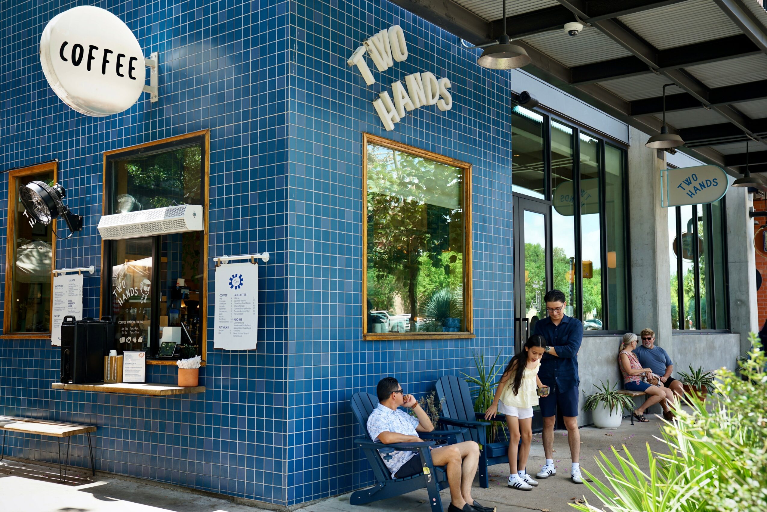 Blue tiled building with signs for coffee and two hands, people sitting and standing outside, large windows, greenery nearby.