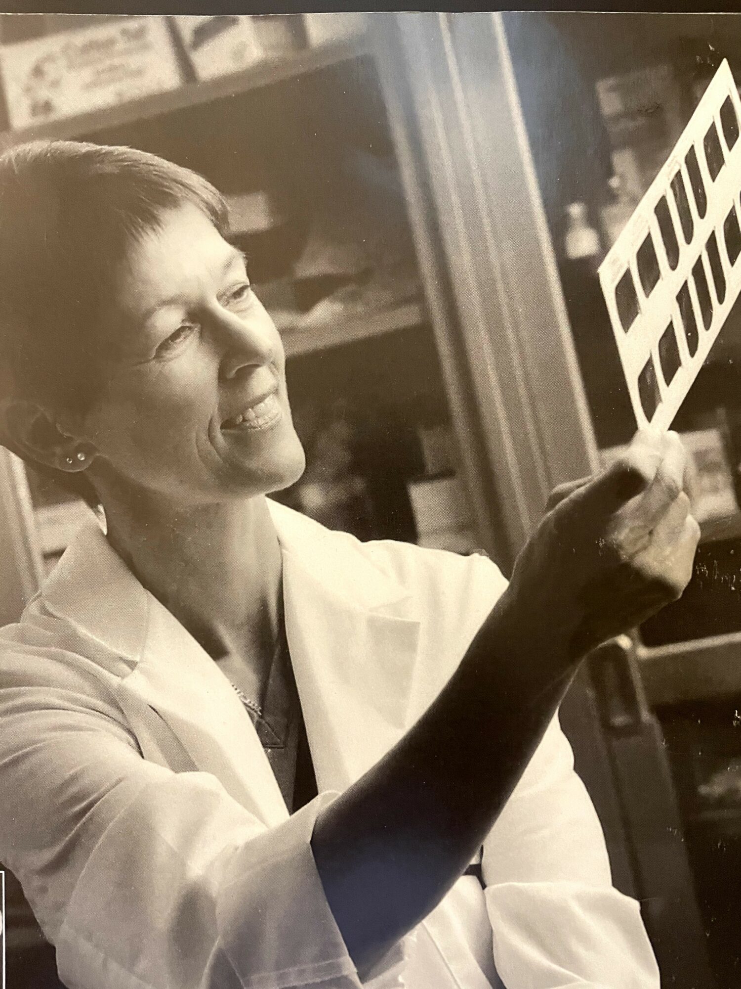Woman in a lab coat holding a film strip, smiling, in a room with shelves and equipment.