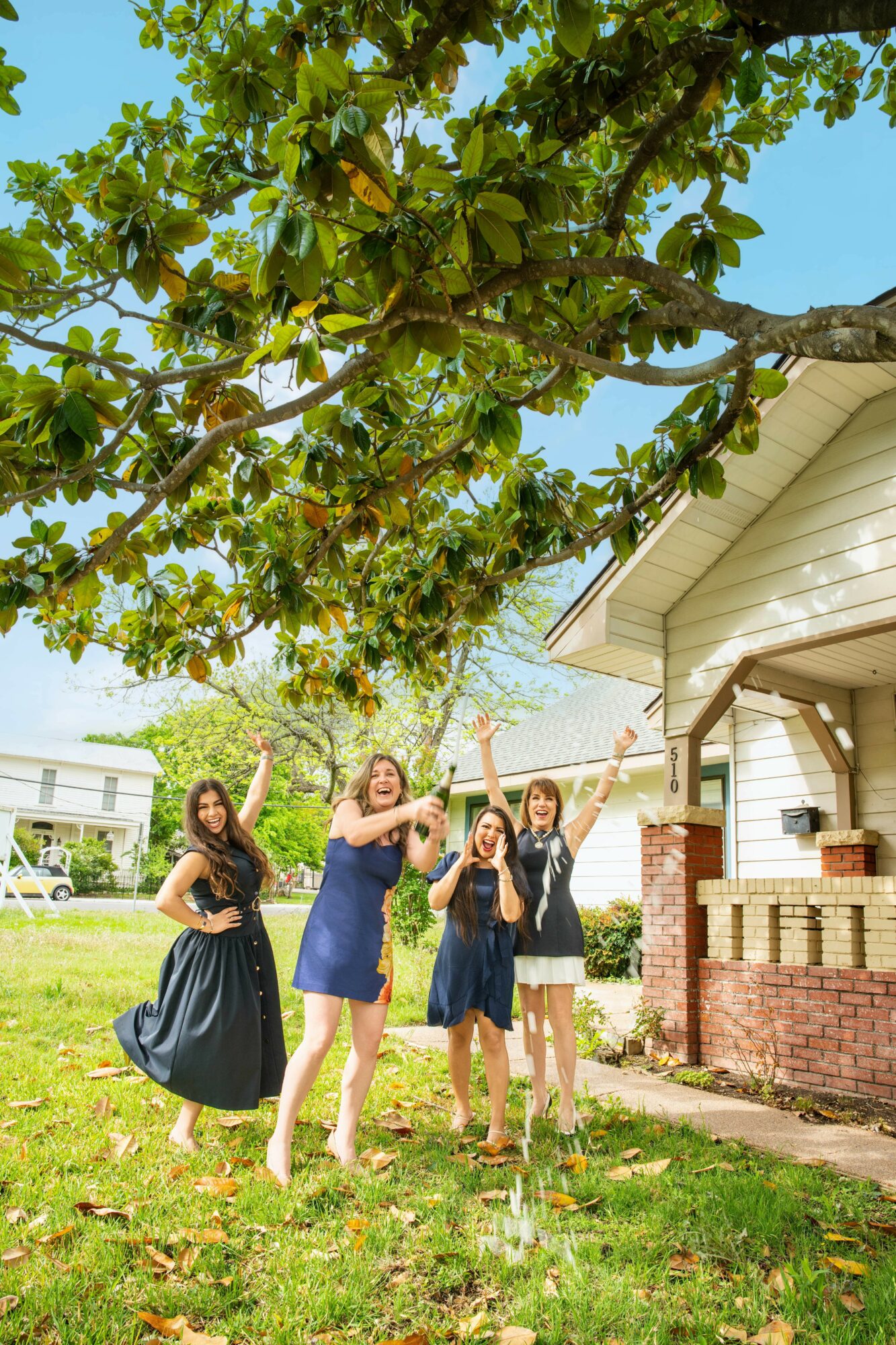 Four girls stand under a large tree in a yard, with a house and a car in the background, sunny day.