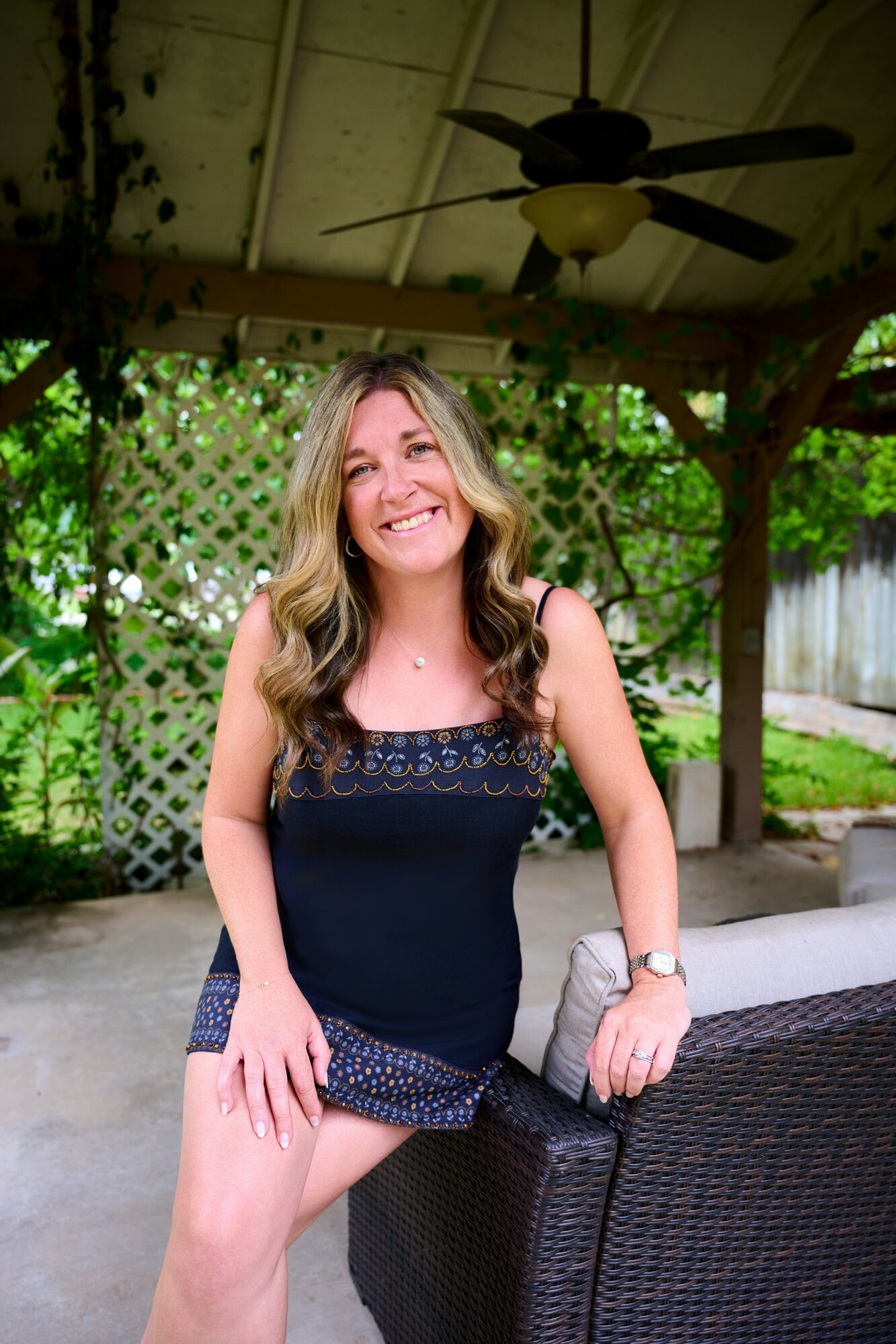 Smiling woman with long hair in a sleeveless navy top, standing outdoors under a pergola with greenery.