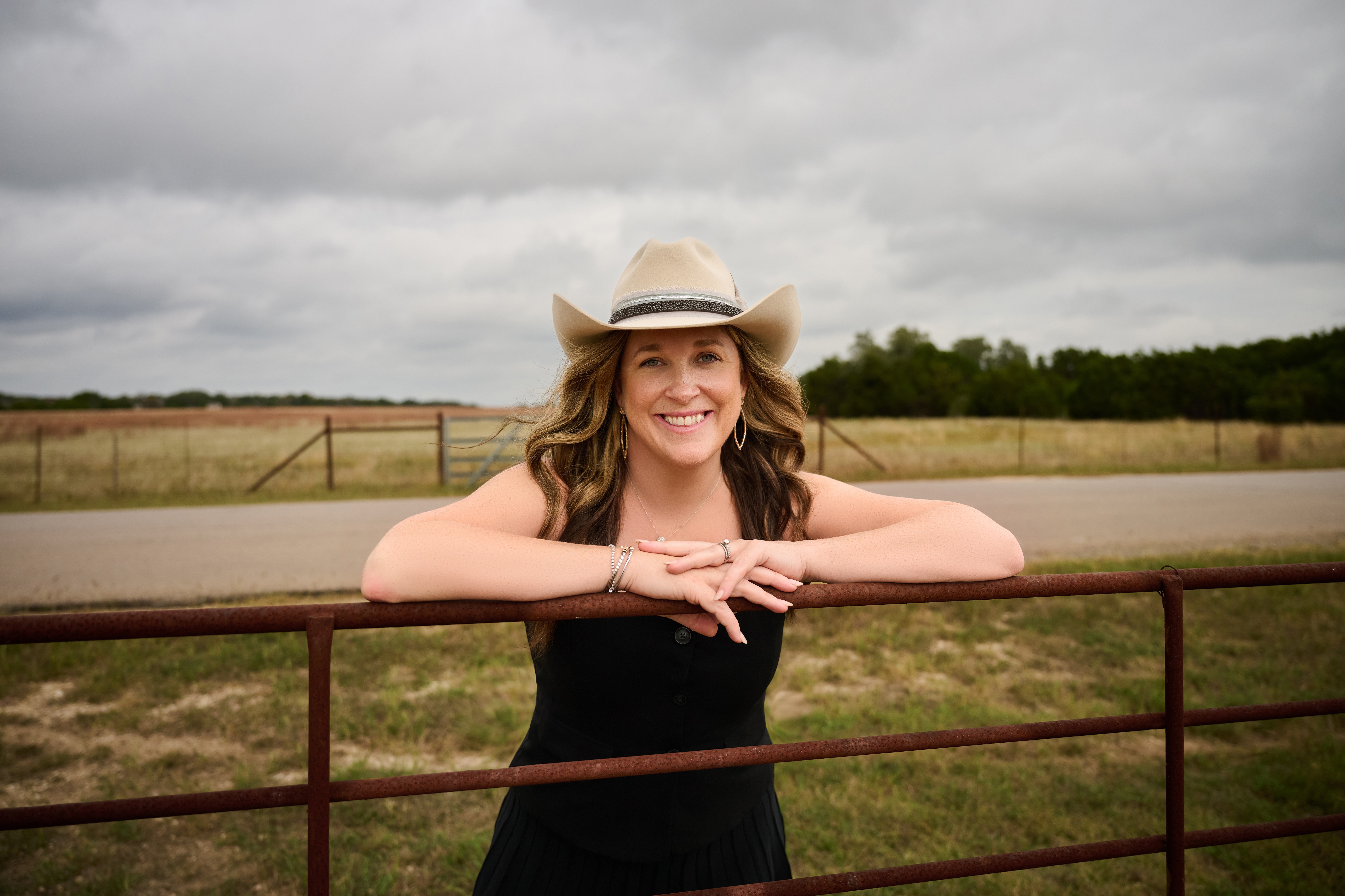 Woman wearing a hat leaning on a fence outdoors, smiling, cloudy sky in background.