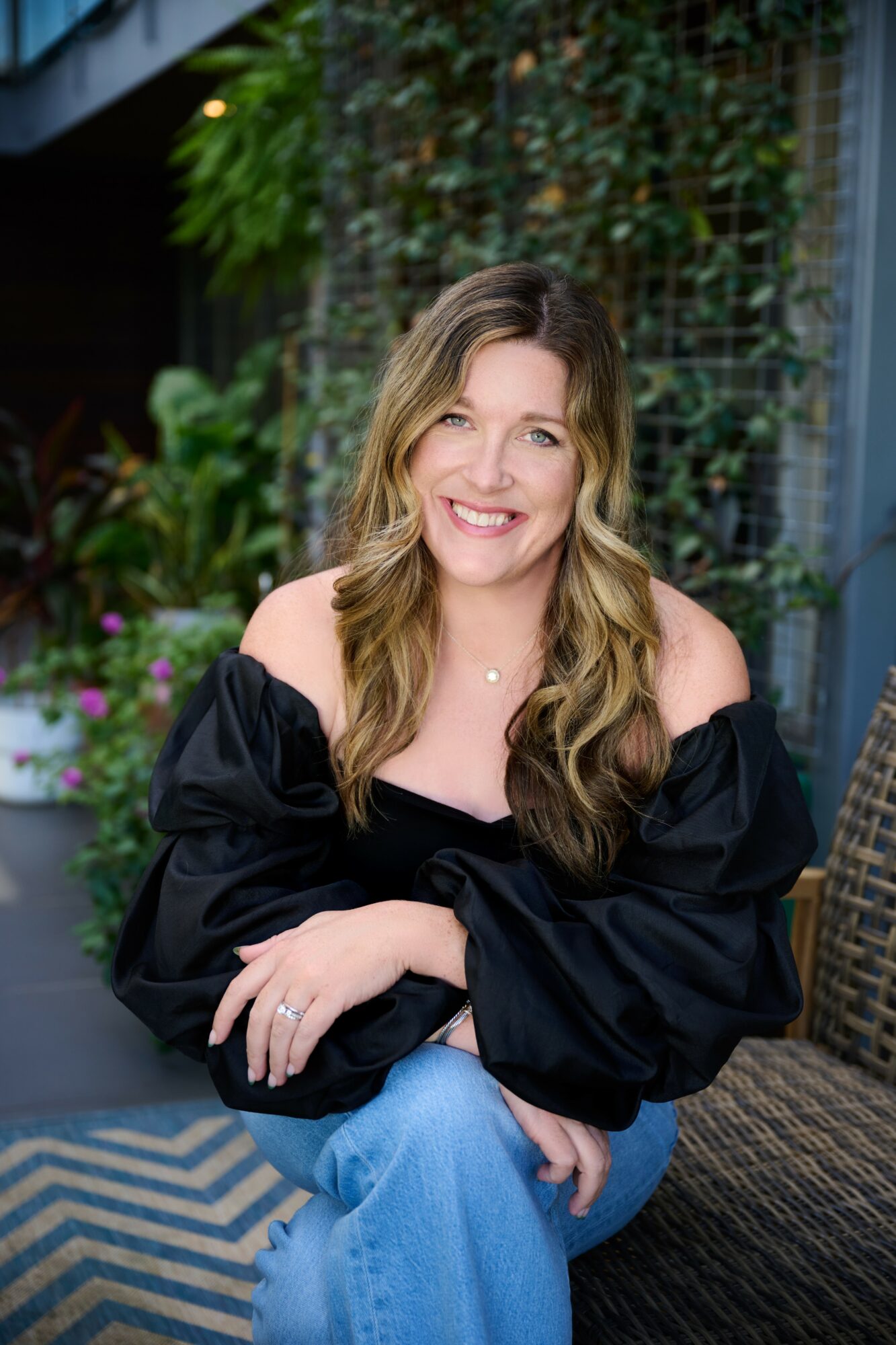 Woman with long wavy hair smiling, sitting outdoors with greenery and a woven chair behind her.