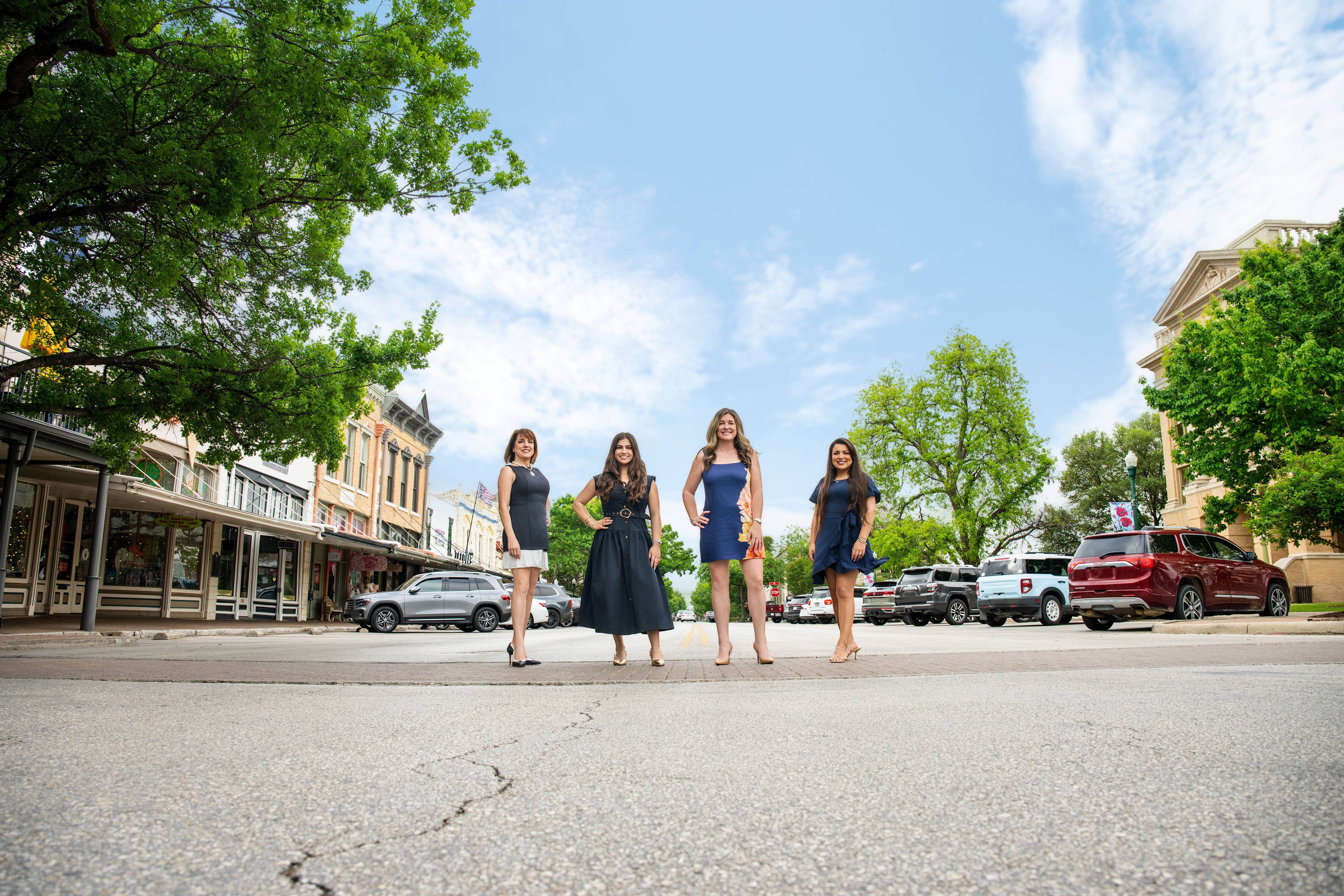 Four women in dresses walking across a street with trees and buildings in the background under a partly cloudy sky.
