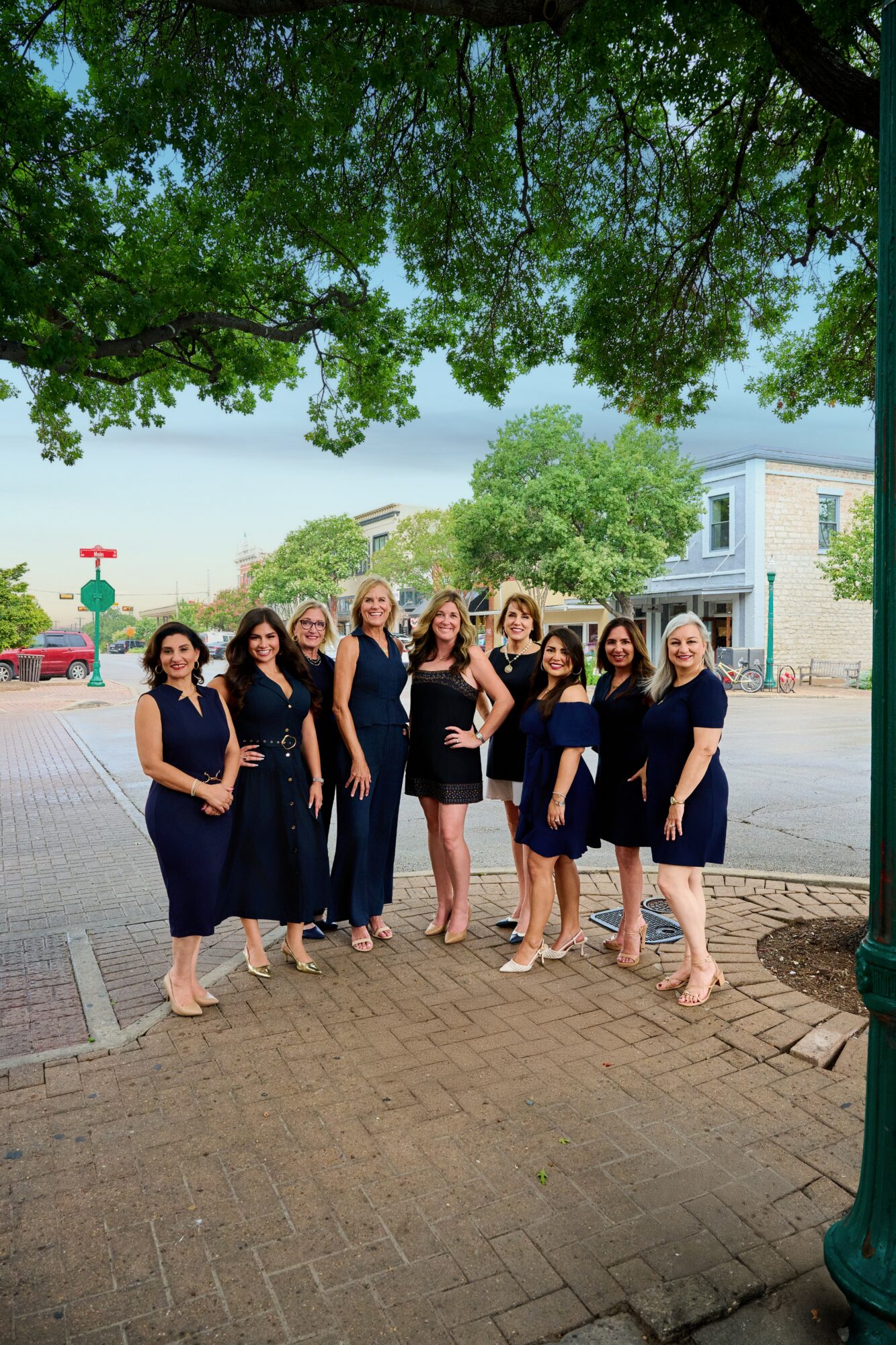 Group of nine women in navy dresses standing outdoors under a tree, smiling, with buildings and a street in background.