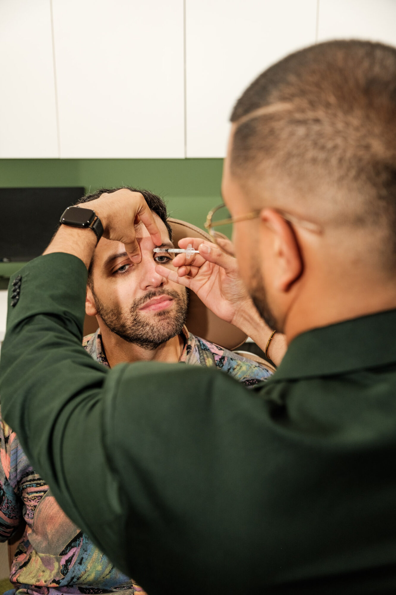 Person receiving makeup application from another person in a room with white cabinets, close-up view.