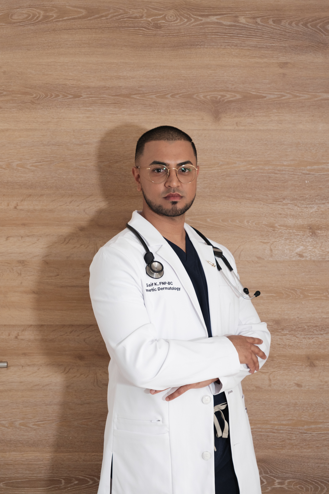Male doctor in white coat with stethoscope, standing against wooden background, arms crossed, looking at camera.
