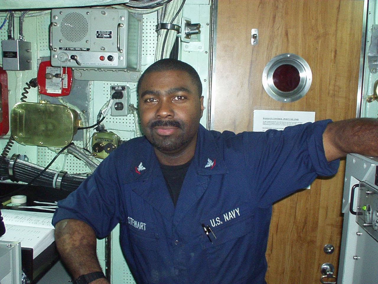 Man in navy uniform with 'U.S. Navy' on chest, standing in a room with equipment, arm extended to the side.