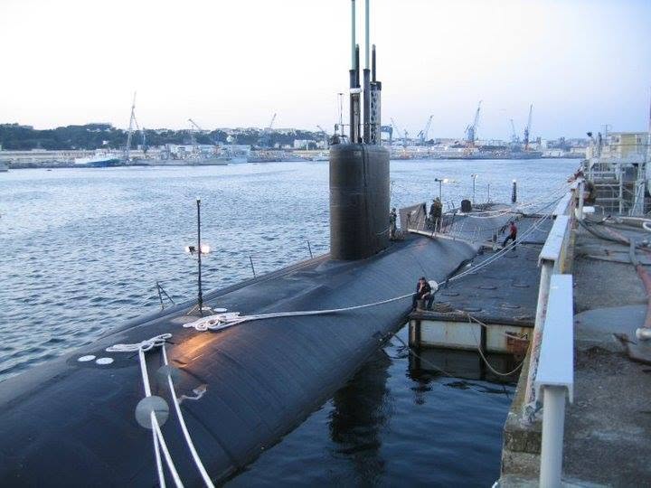 Submarine docked at a harbor with cranes and ships in the background, moored alongside a pier with ropes and safety barriers.