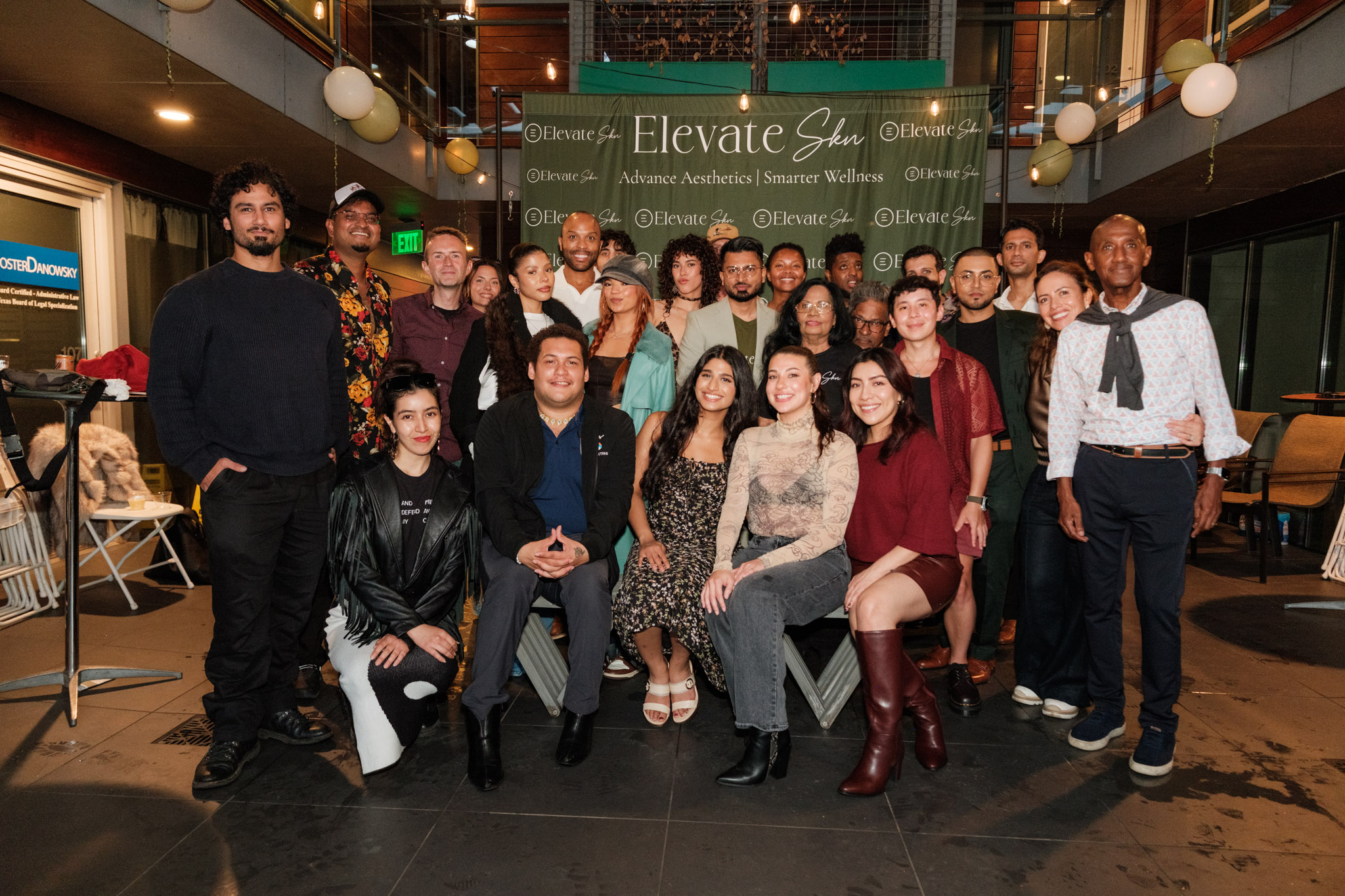 Group of people posing indoors in front of a green banner, some seated and some standing, with warm lighting.