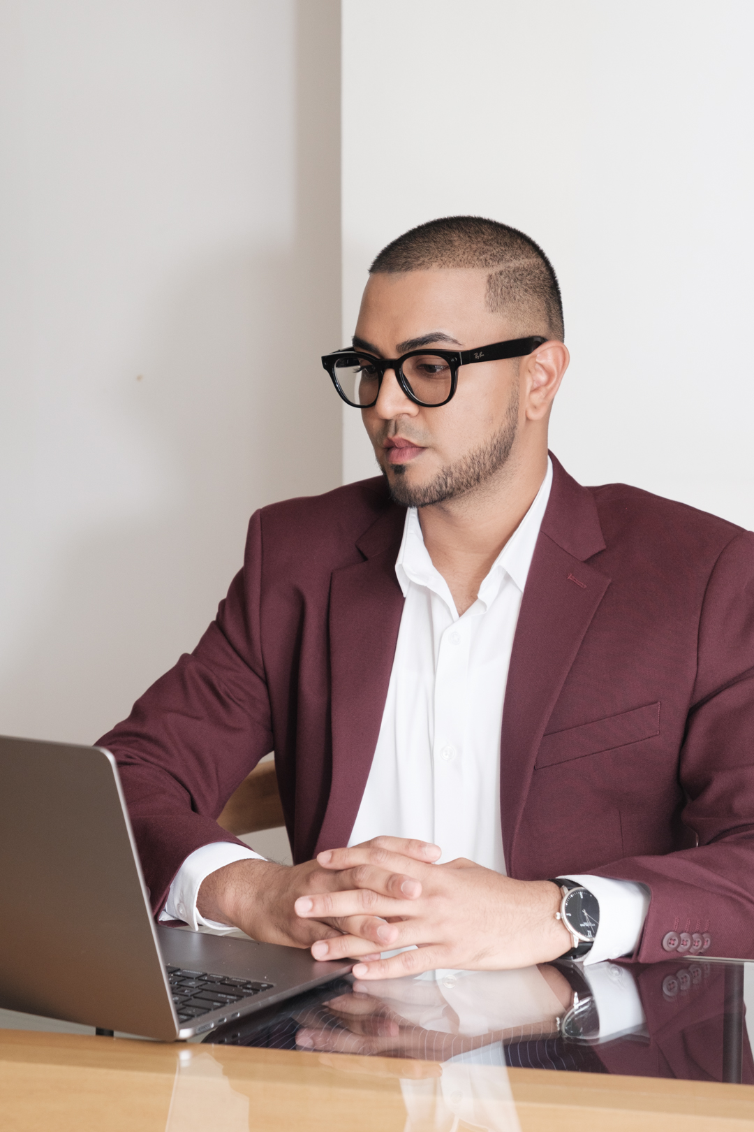 Man with glasses and a beard wearing a maroon blazer and white shirt, sitting at a desk with a laptop.