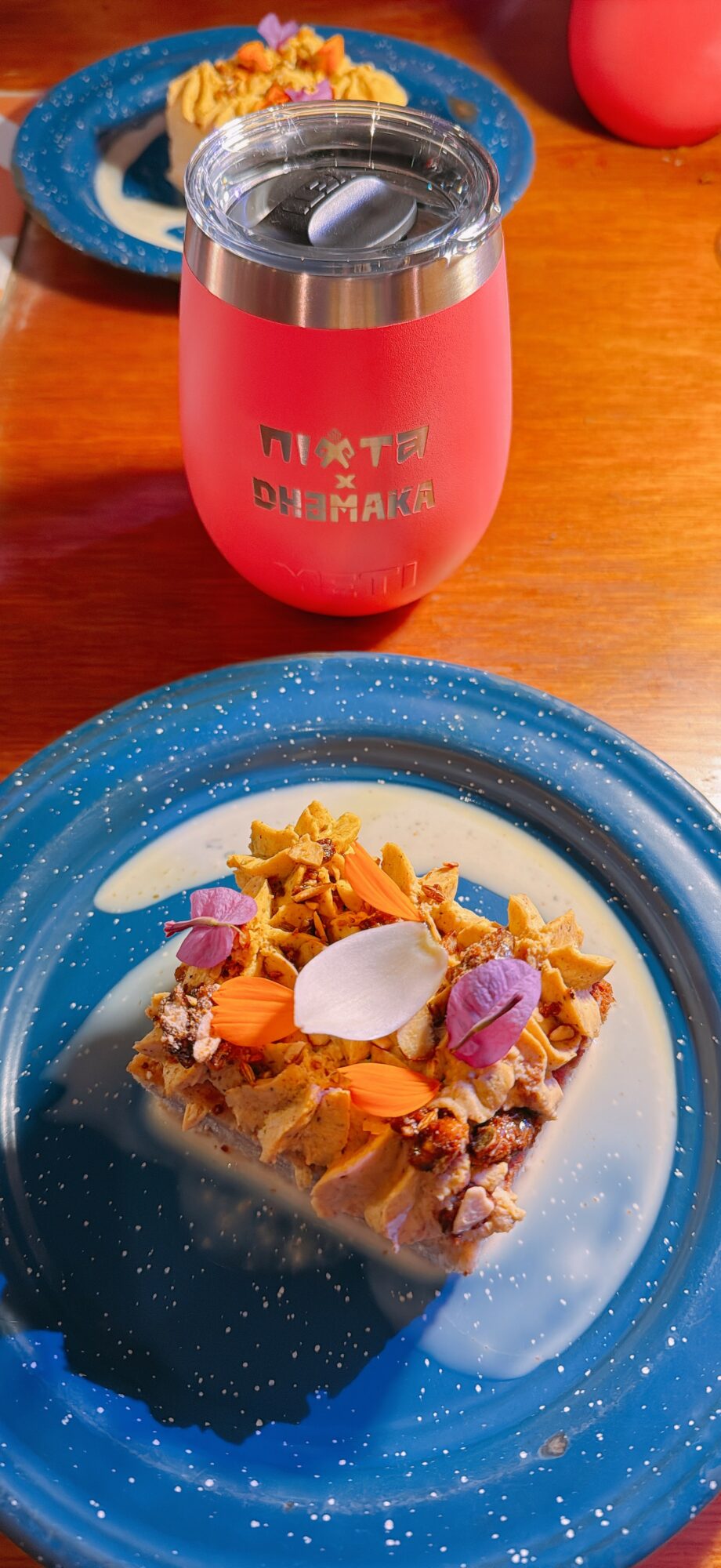 A slice of cake with colorful toppings on a blue plate, and a pink tumbler with a silver lid on a wooden table.