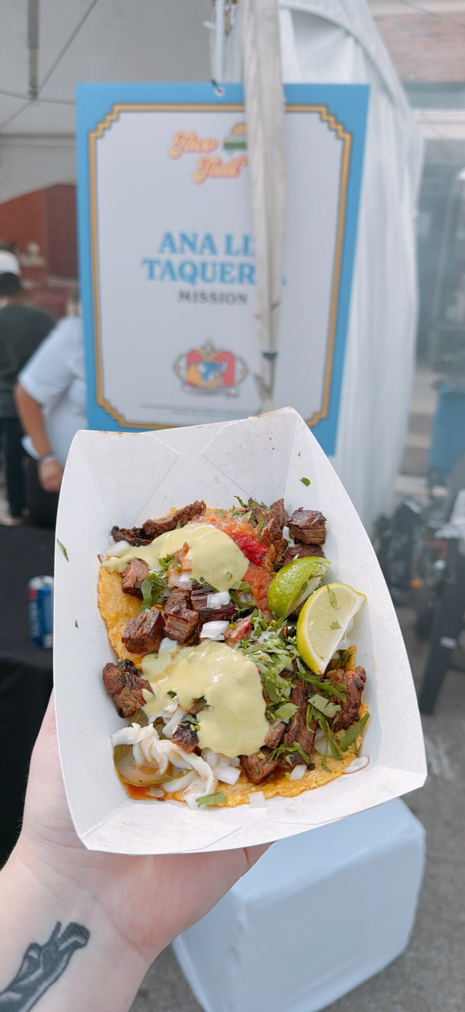 Hand holding a tray of food with meat, cheese, lime, and vegetables in front of a sign with text and logo.