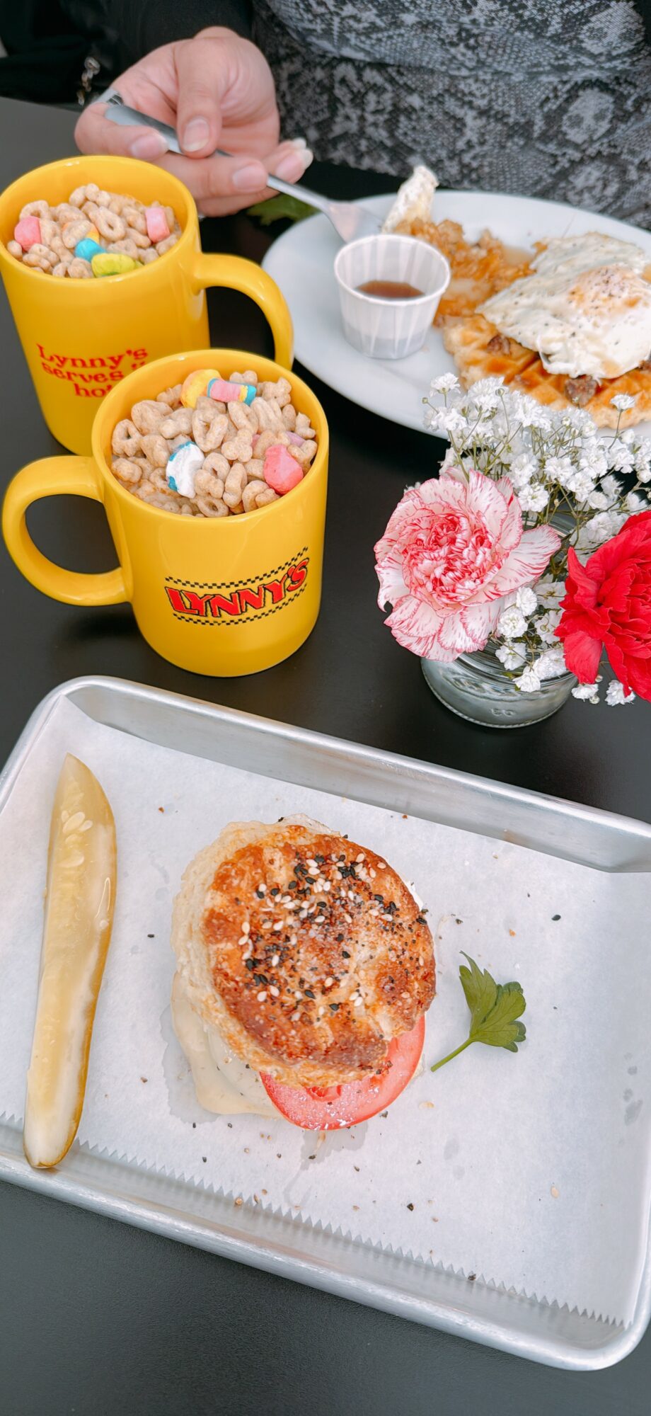Two yellow mugs filled with colorful cereal, a plate with a burger, fries, and a pickle, flowers in a vase, and a cup of coffee.