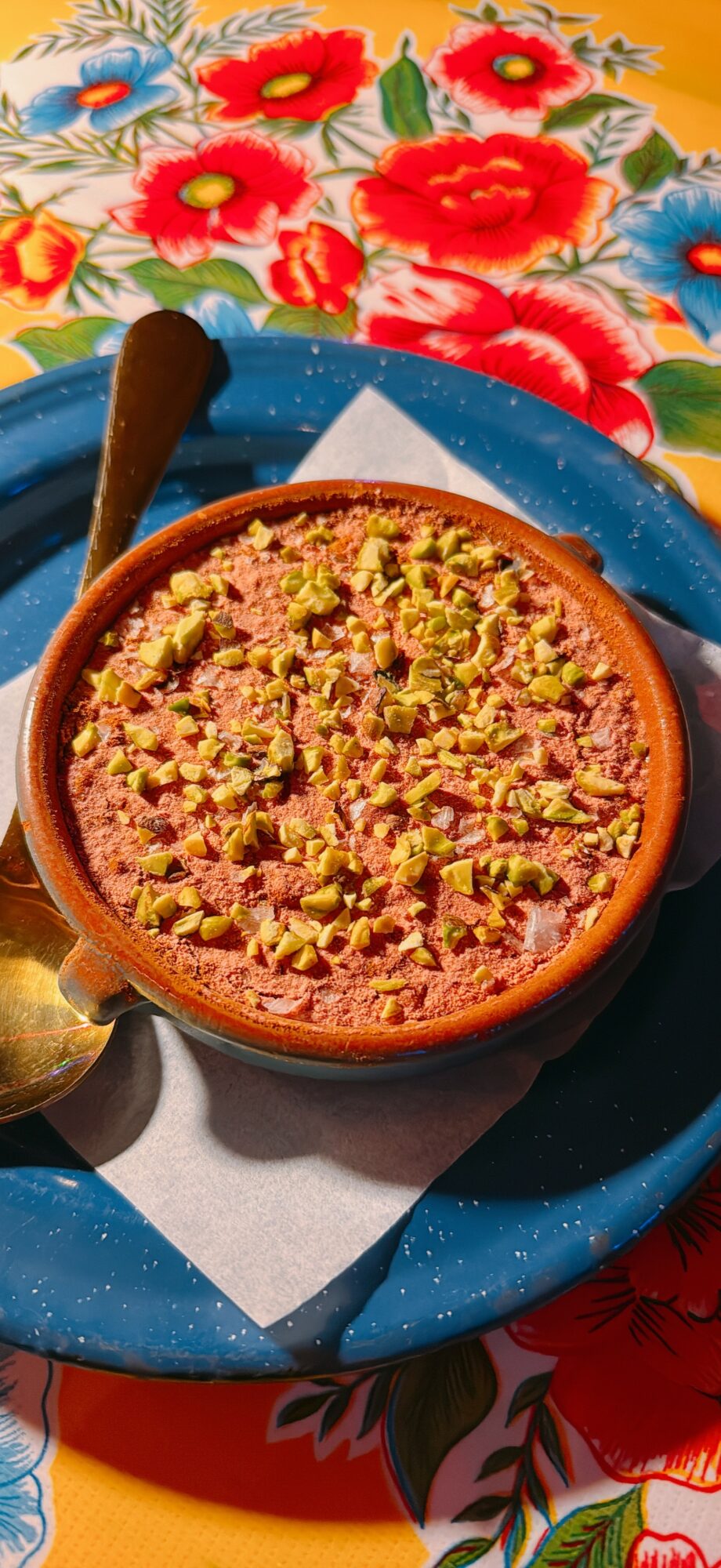 Baked dessert topped with chopped pistachios on a blue plate, with colorful floral tablecloth in background.