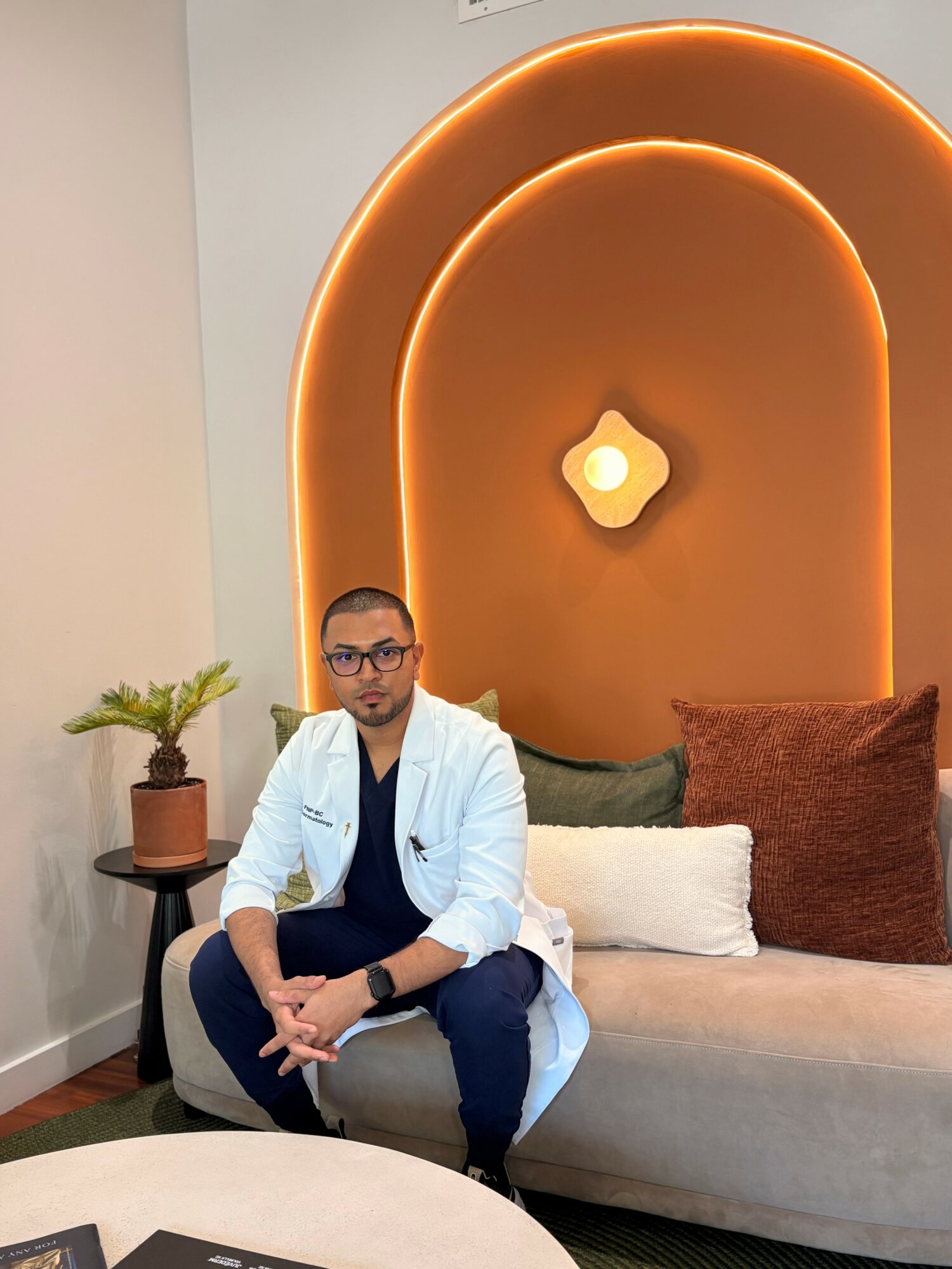 Person sitting on a sofa in a room with decorative wall lighting, wearing a white coat and glasses.