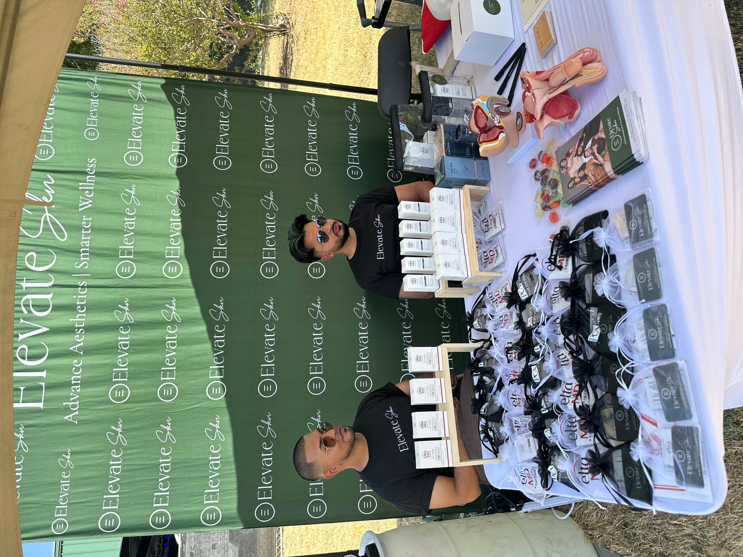 Table with various products and two women standing behind it, in front of a green backdrop with text. Items include bottles, boxes, and packaged goods.