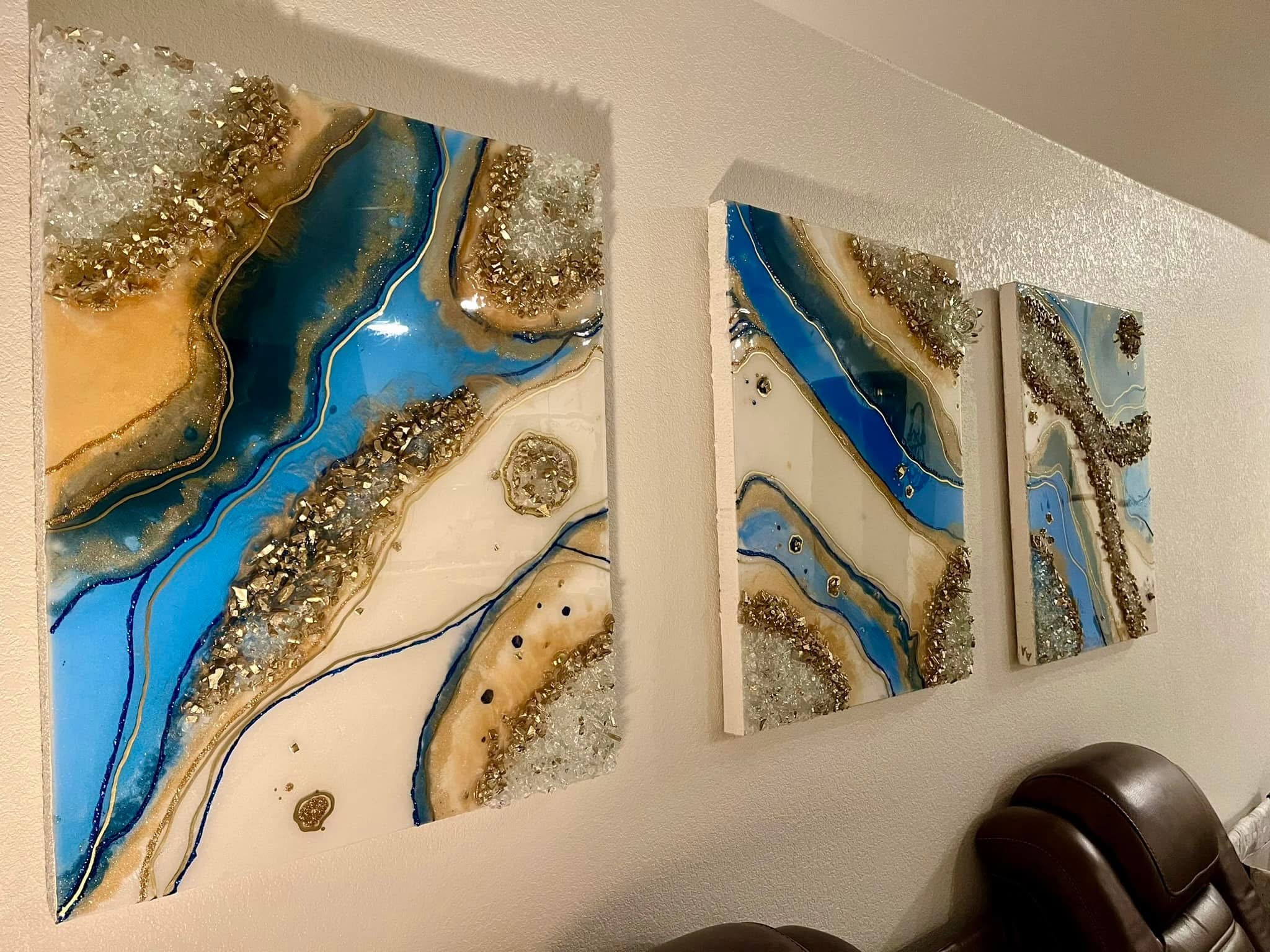Three abstract paintings with blue, gold, and white colors hang on a wall, with a dark chair partially visible in the corner.