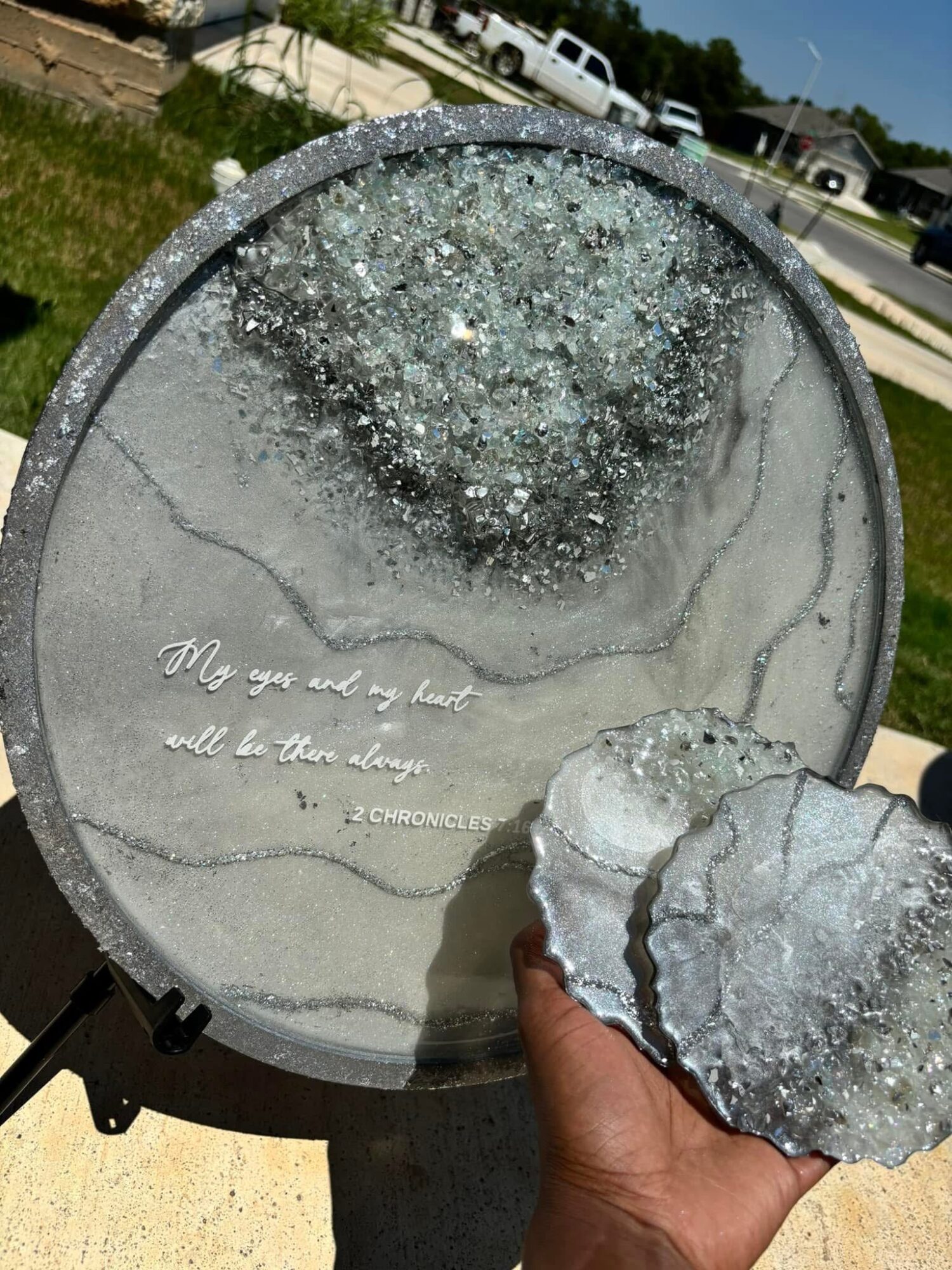 Person holding a broken glass piece in front of a decorative round glass panel with a quote, outdoors on a sunny day.