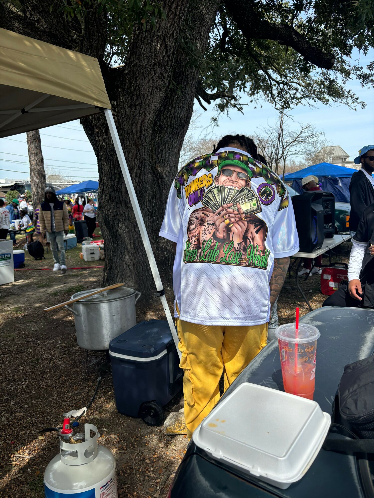 Person wearing a colorful shirt with a tiger design, yellow pants, and a mask, standing outdoors near trees and tables.