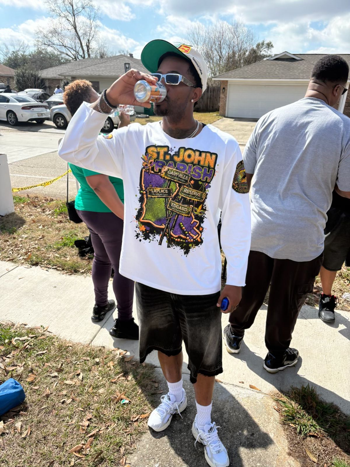 Person wearing sunglasses, a white T-shirt with a graphic, black shorts, and white sneakers, drinking from a bottle outdoors.