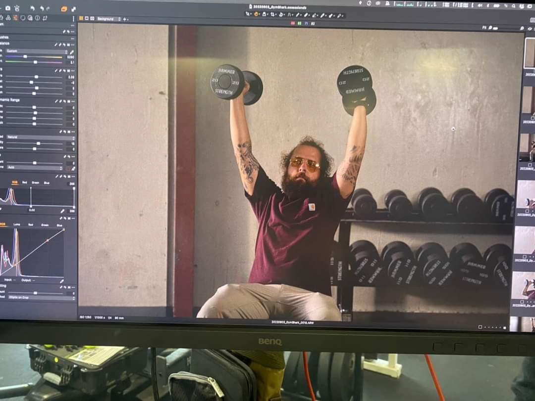 Person with a beard and tattoos lifting dumbbells in a gym, sitting on a bench, with a rack of weights behind.