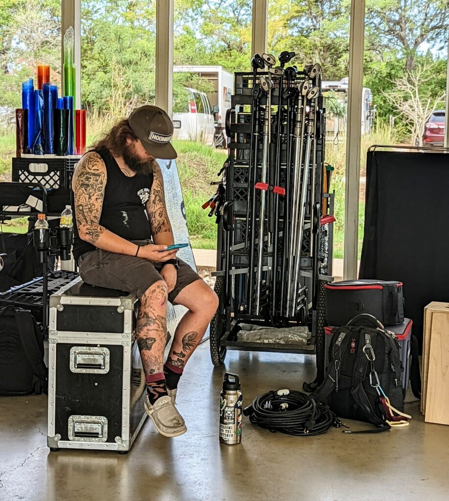 Woman with tattoos sitting on a black box, looking at her phone, in a room with large windows and fitness equipment.