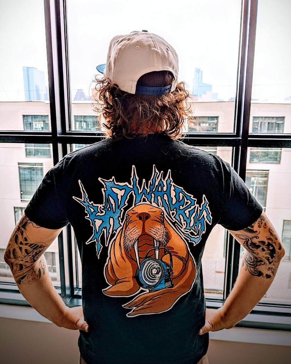 Person with curly hair wearing a cap and a graphic T-shirt, standing in front of large windows with city buildings outside.