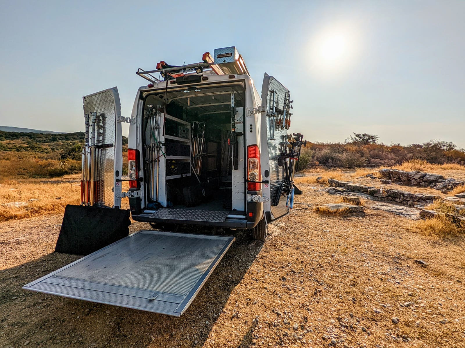 Back of a service van with open doors and equipment, on a dry, open landscape with a bright sun.