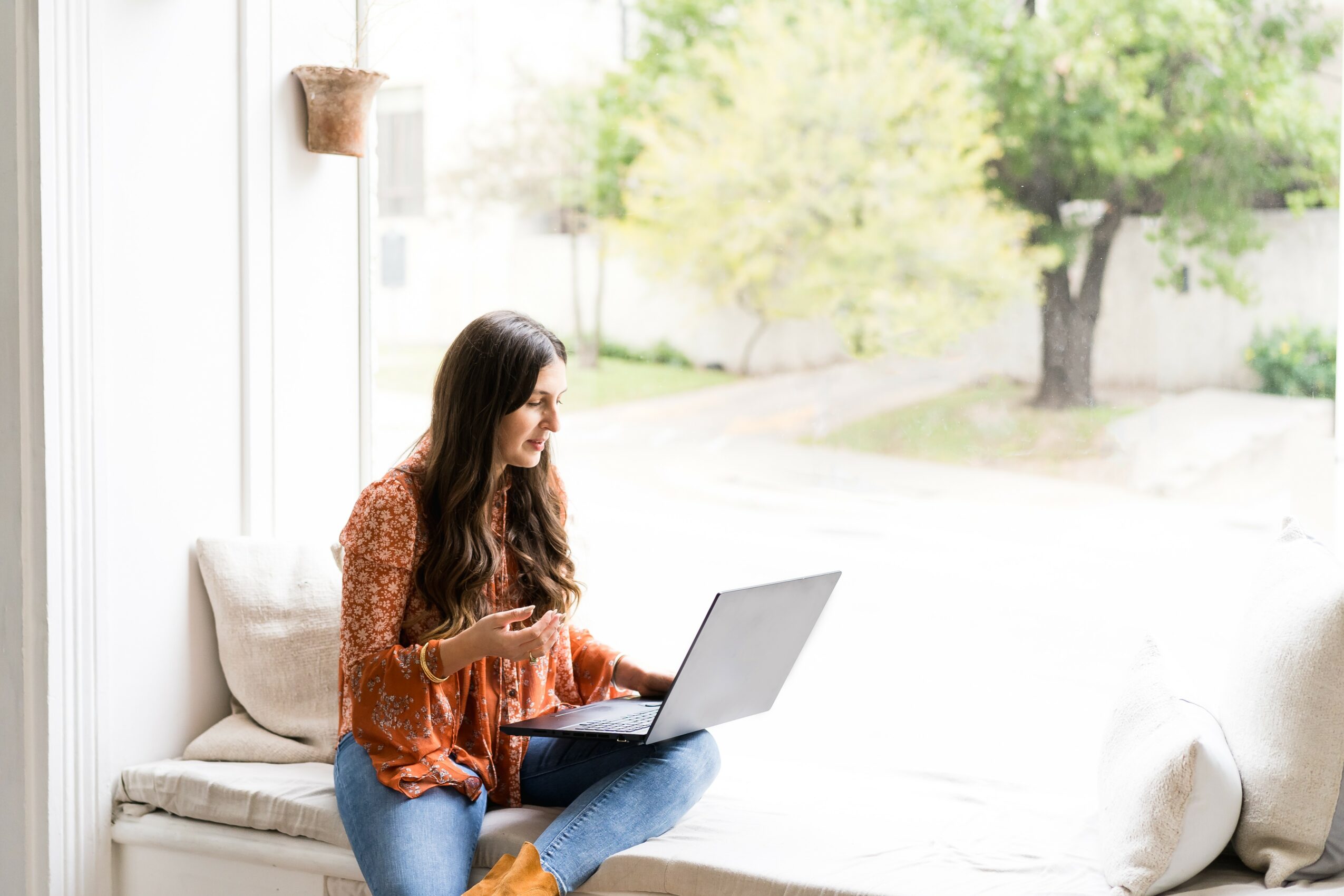 Woman sitting on a window seat using a laptop outdoors, with trees and a yard visible in the background.