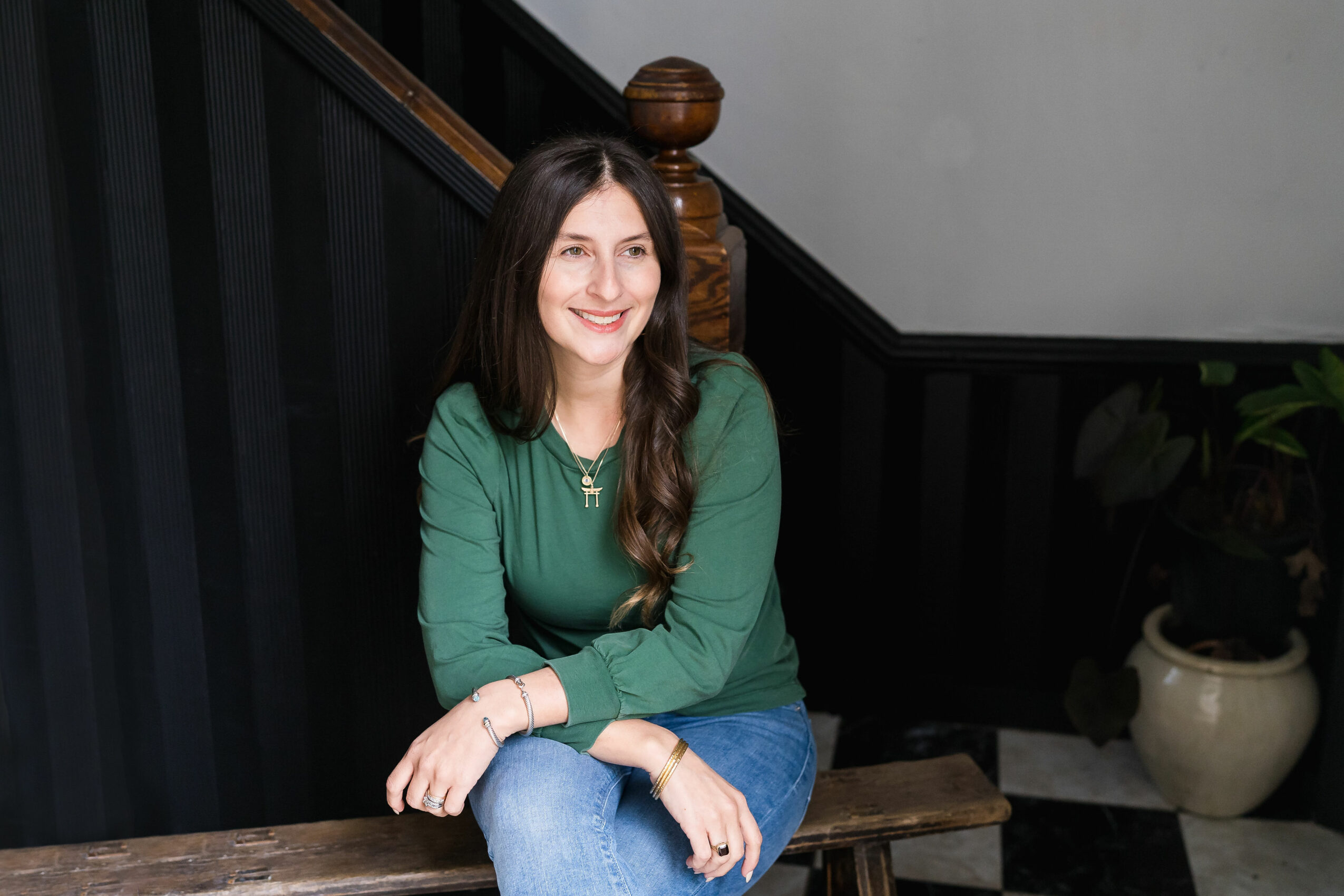 Young woman with long dark hair smiling, wearing a green top and jeans, sitting indoors near a staircase.