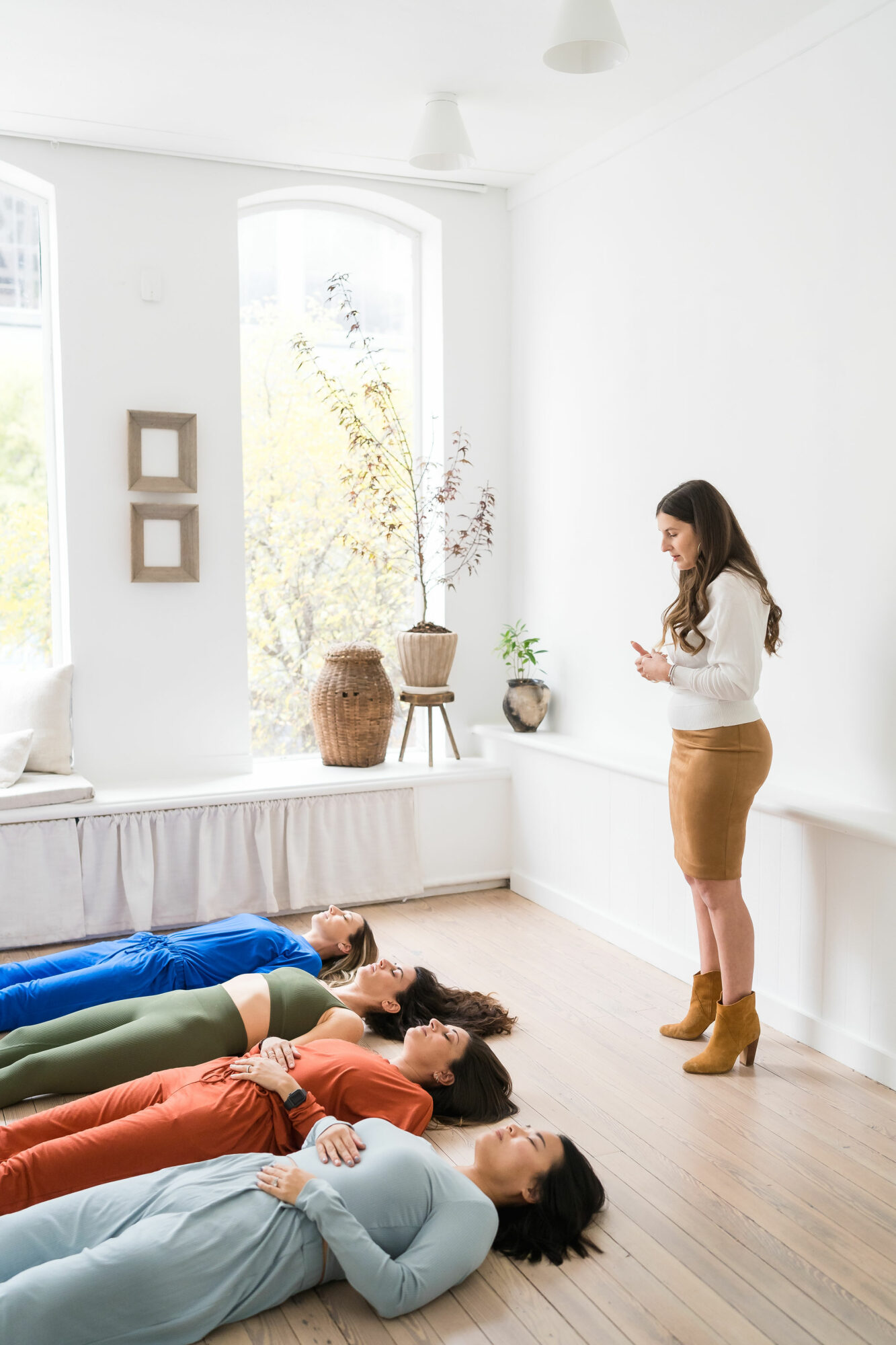 Four people lie on the floor with heads together, a woman stands nearby in a bright room with large windows and decor.