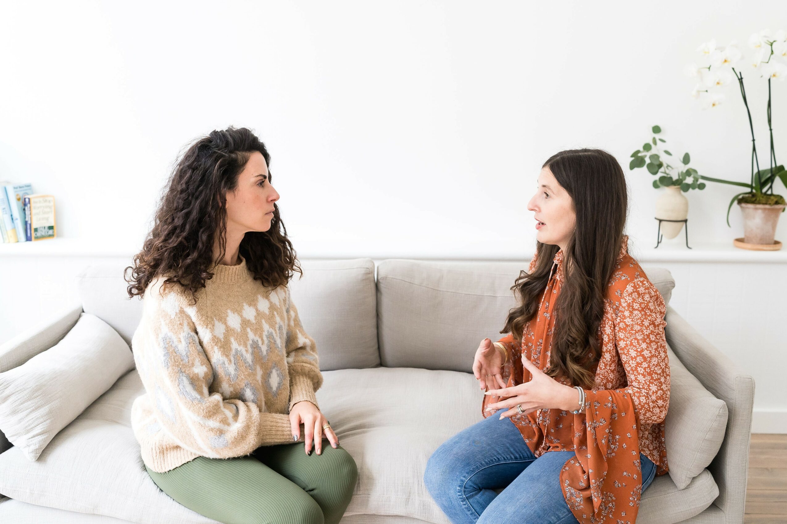 Two women sit on a sofa, engaging in conversation in a bright room with plants and books, one with curly hair, the other with straight hair.