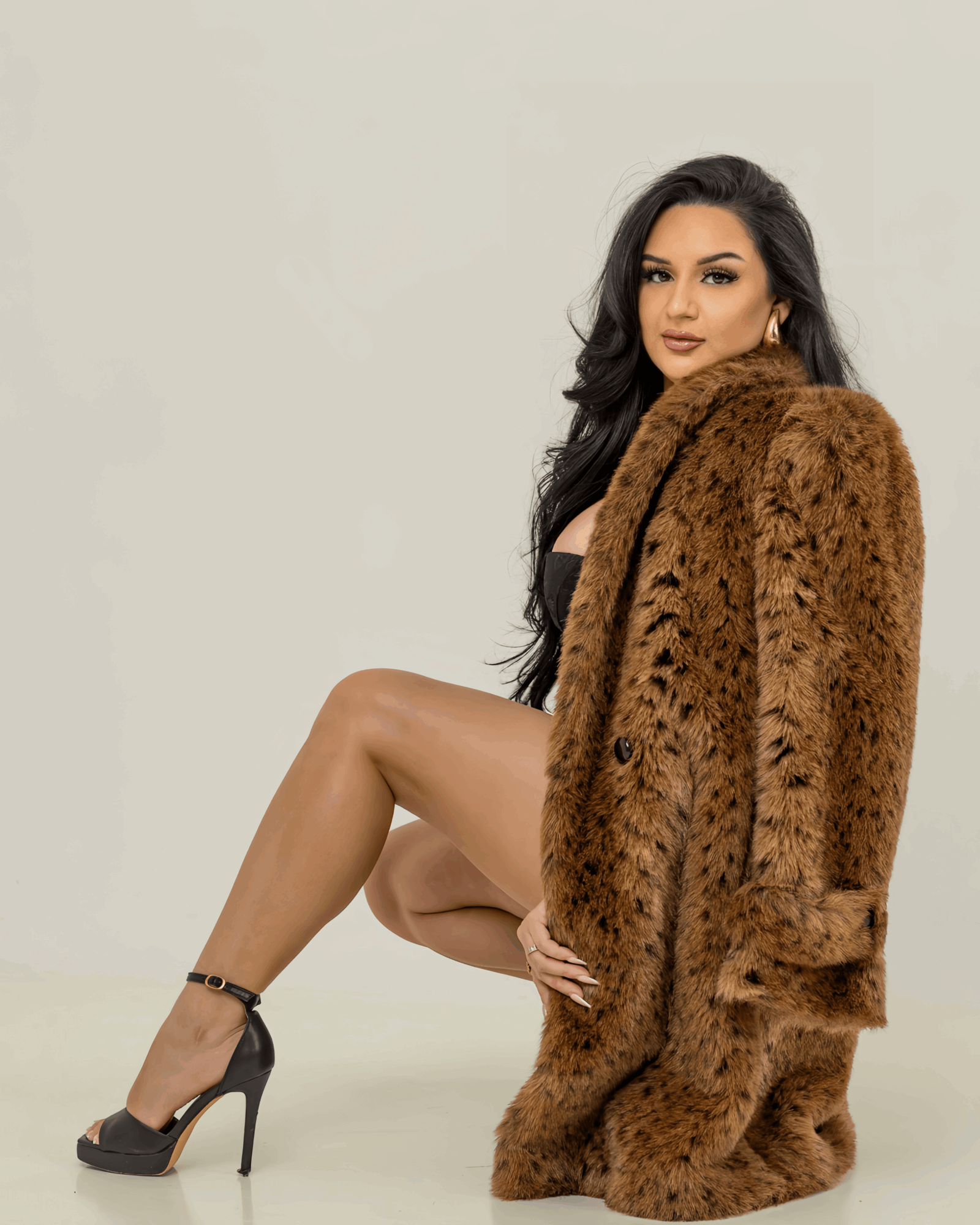 Woman with long dark hair wearing high heels and a large brown fur coat, sitting with one knee up, against a plain background.
